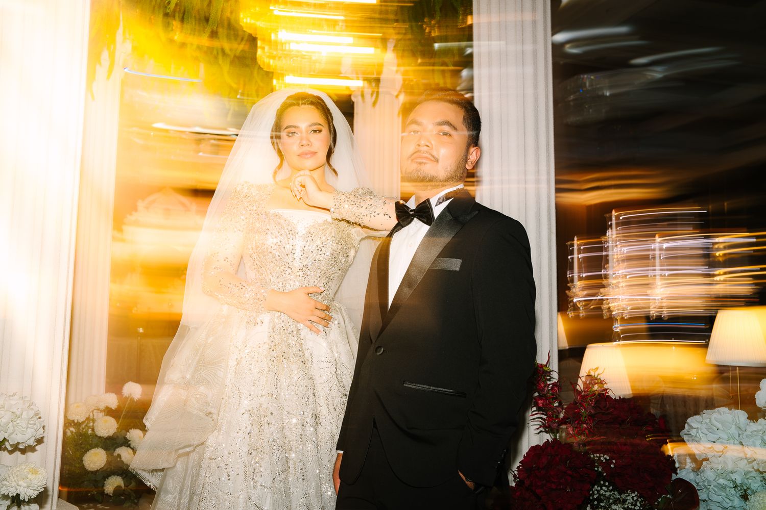 Bride and groom in elegant wedding attire posing with golden light flare effect.