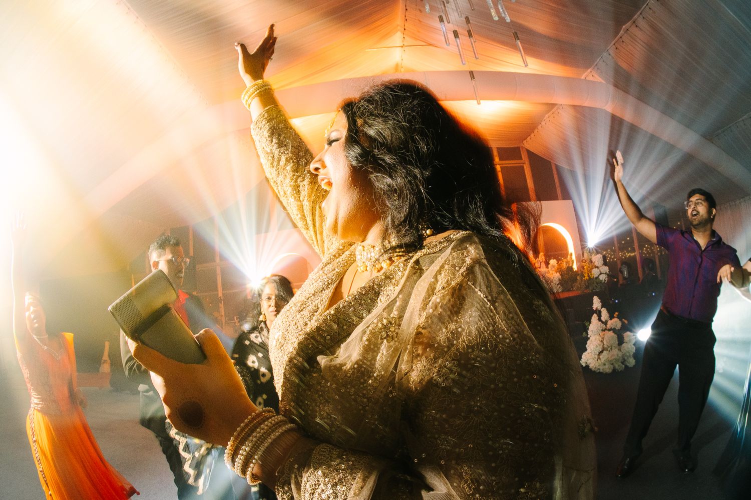 Woman in gold embroidered lehenga dancing joyfully under bright lights at a vibrant wedding reception.