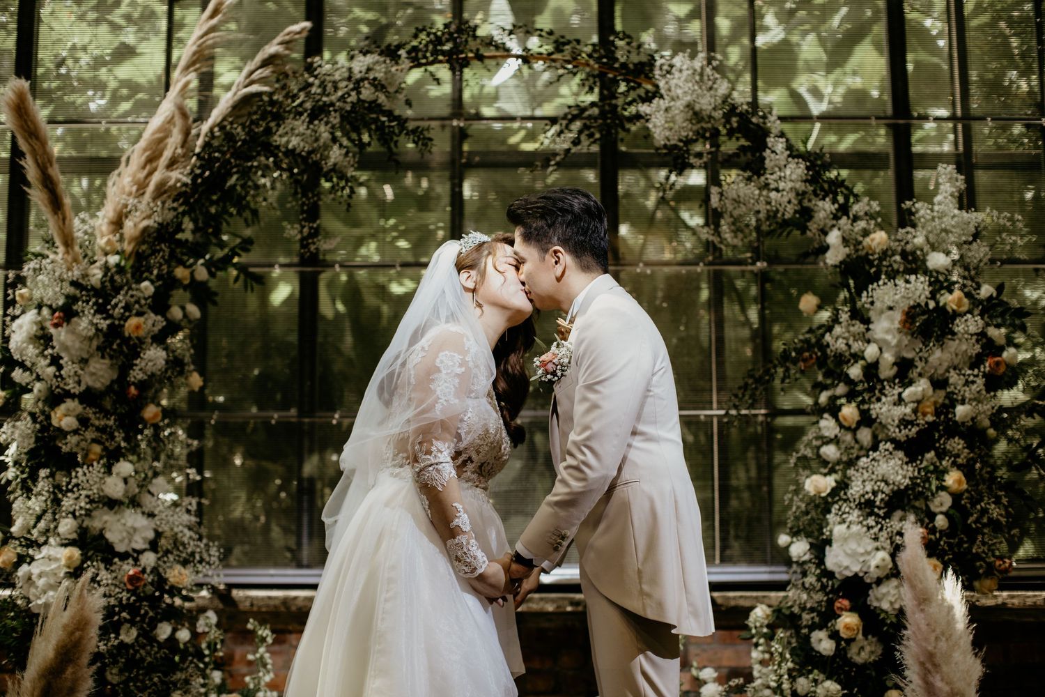 Bride and groom share first kiss under stunning floral arch adorned with pampas grass and white blooms.