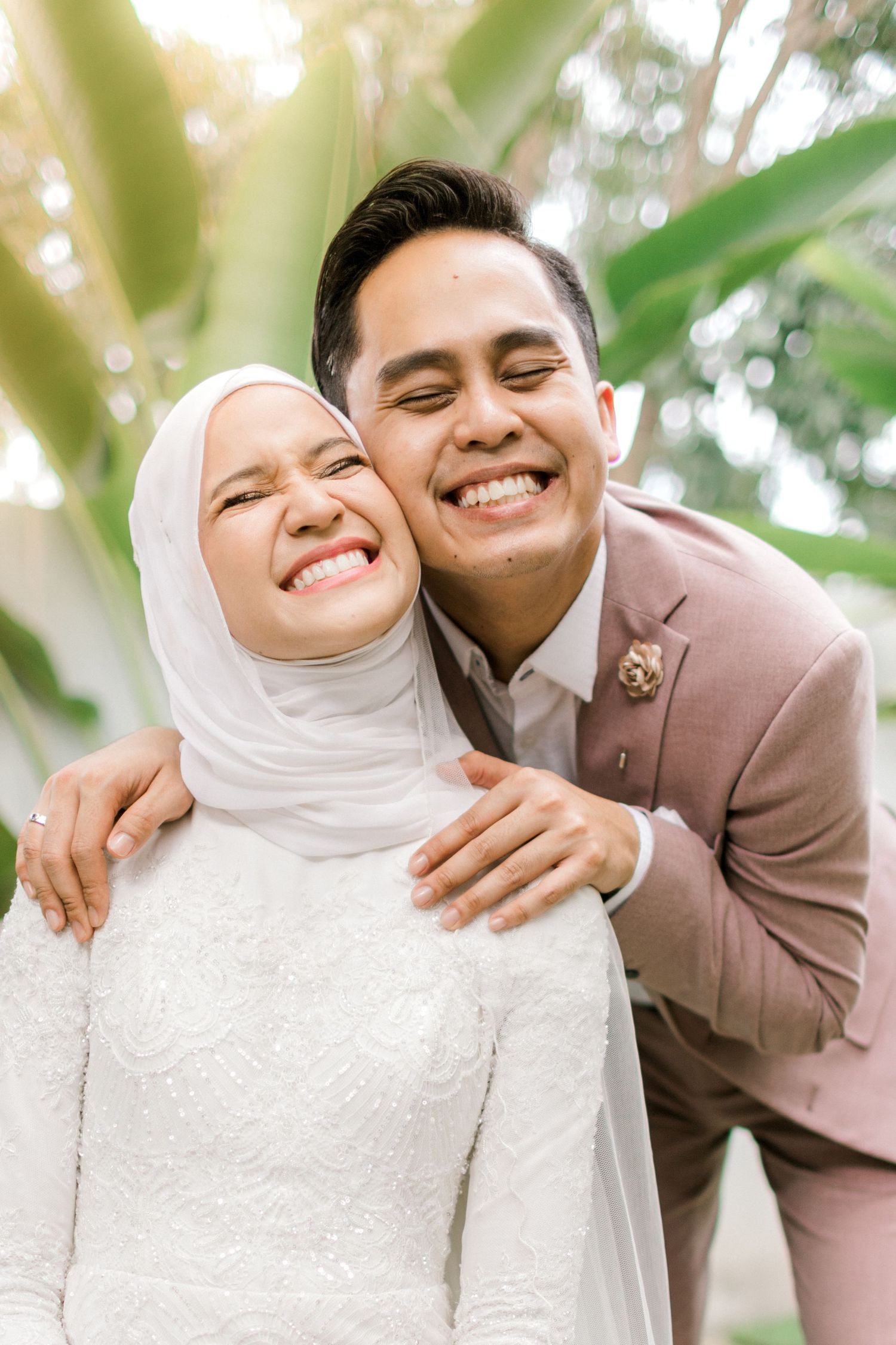 A joyful Malaysian couple in wedding attire smiling outdoors, bride in white hijab and groom in pink suit embracing.