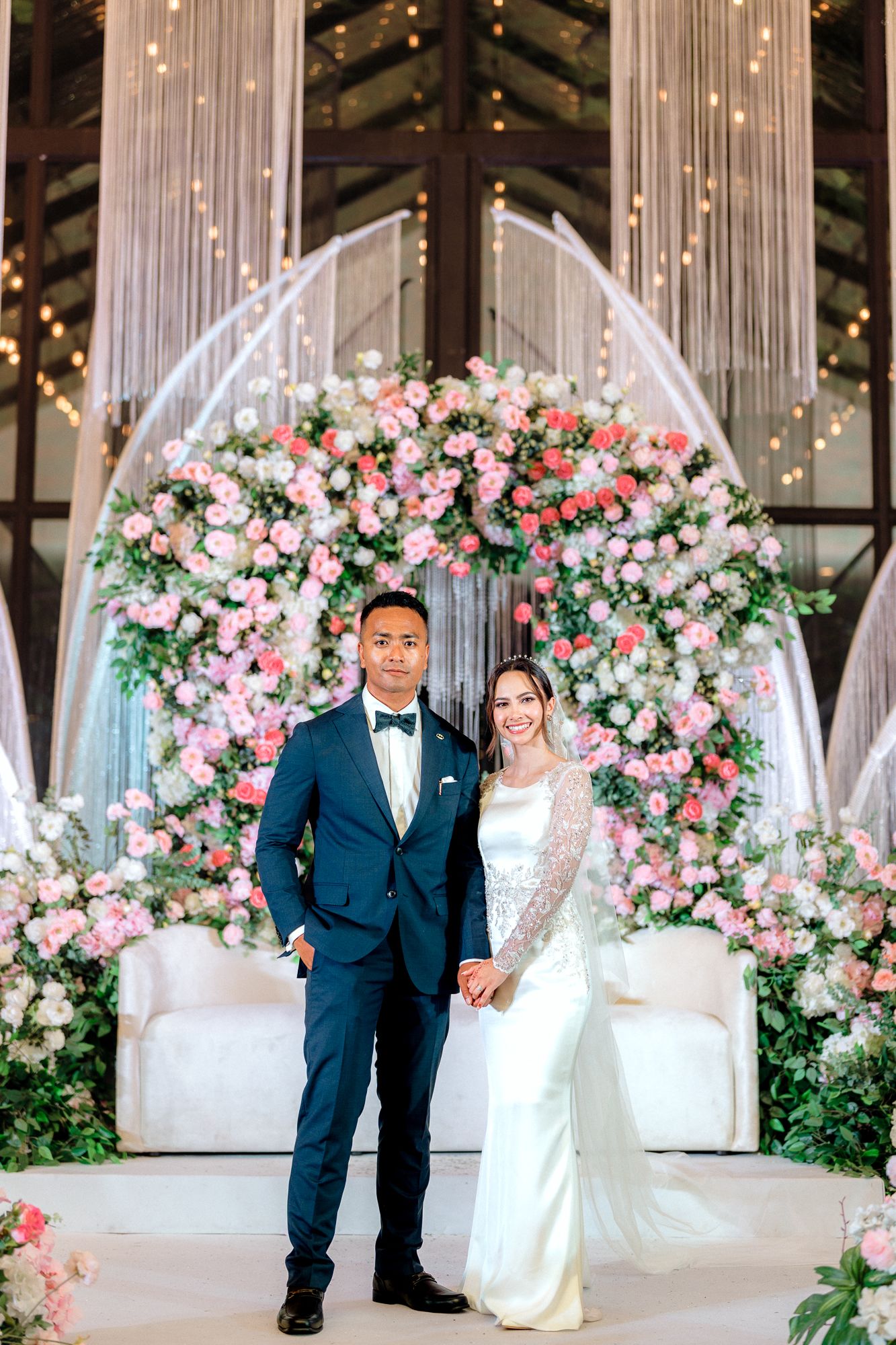 Couple posing before large pink floral arch at elegant indoor wedding reception venue.