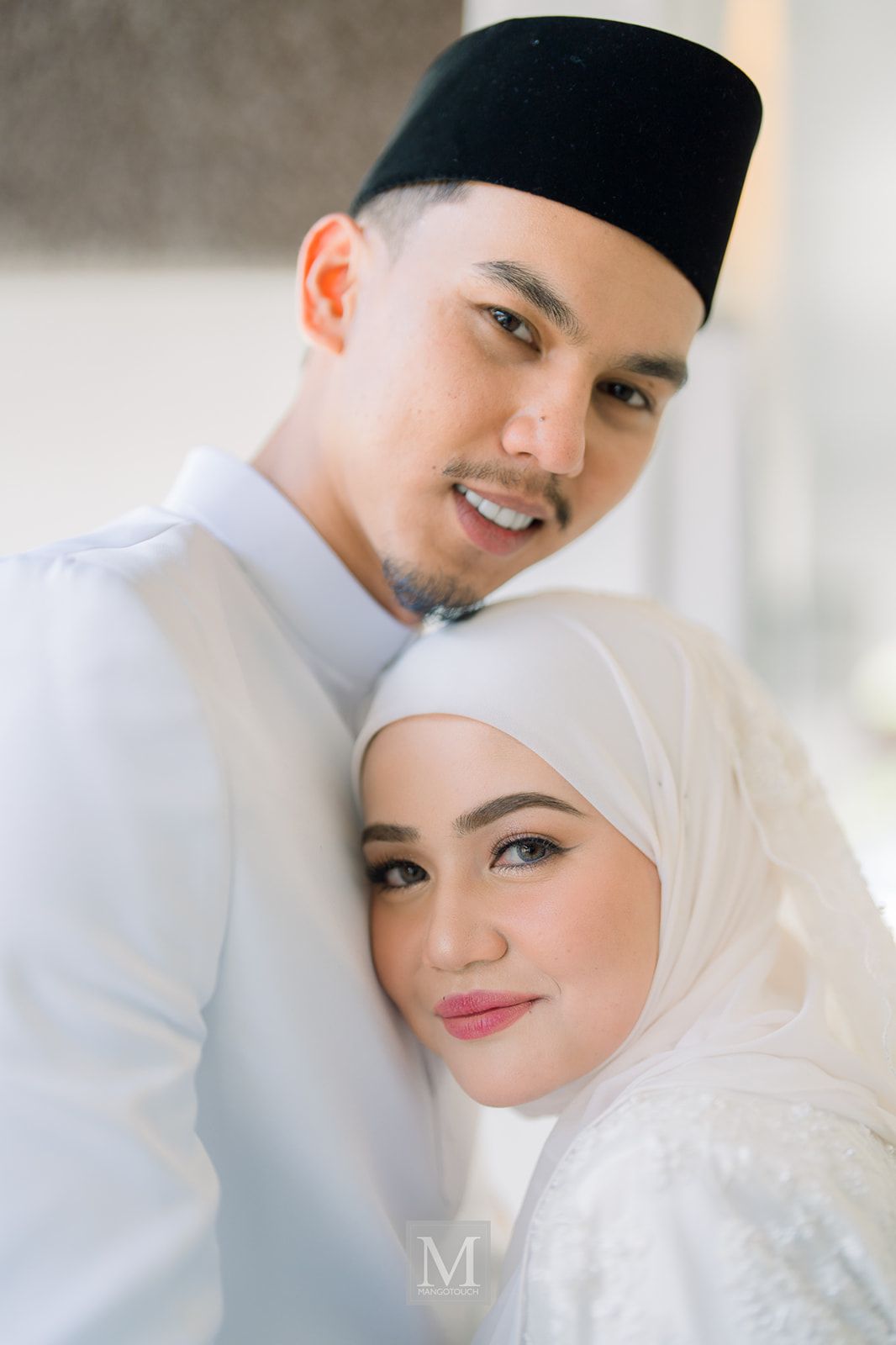 Malaysian couple in white traditional attire posing together, bride wearing hijab, groom in songkok hat.
