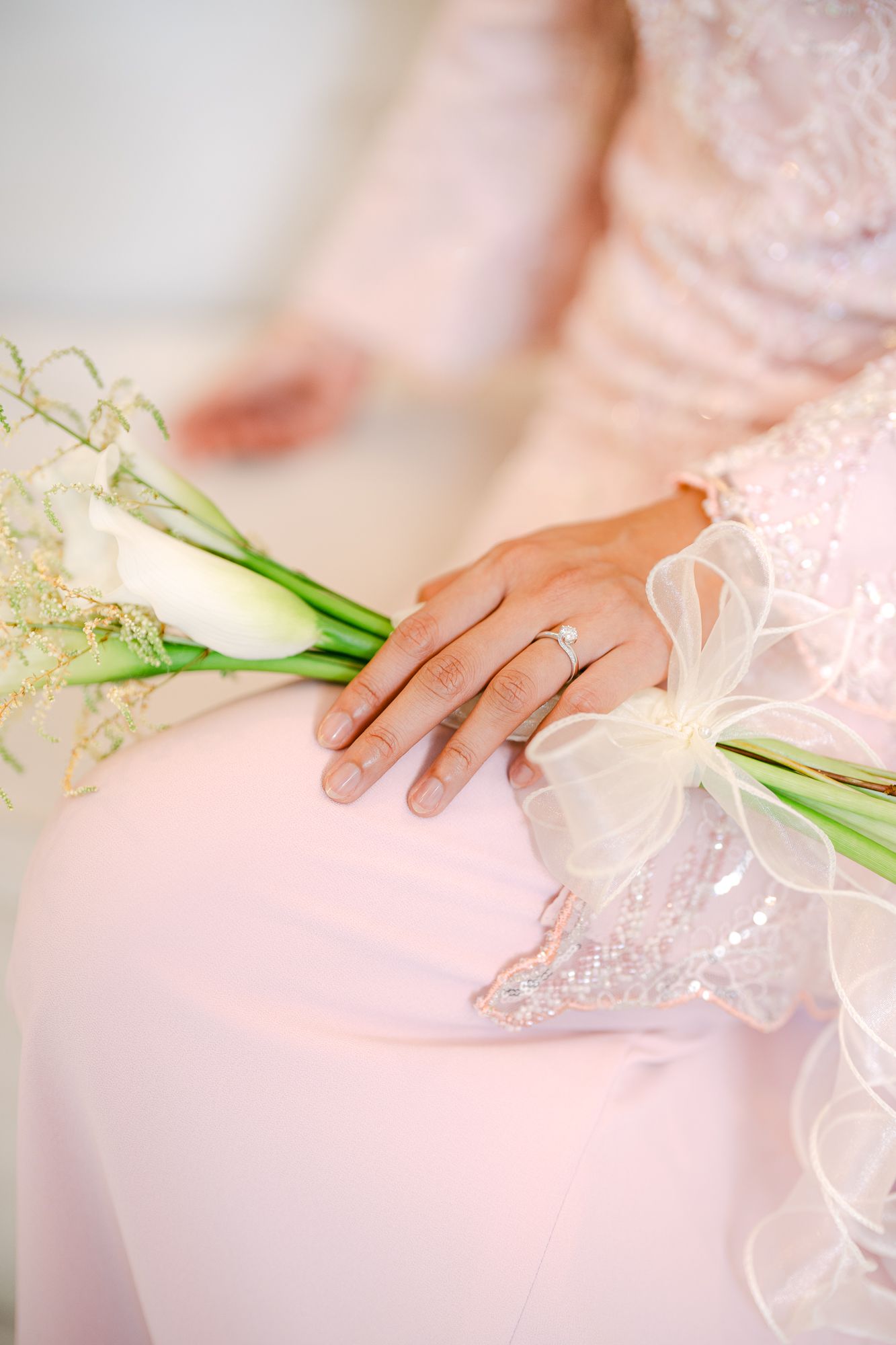 Bride in pink gown holding white calla lily bouquet with diamond engagement ring visible.