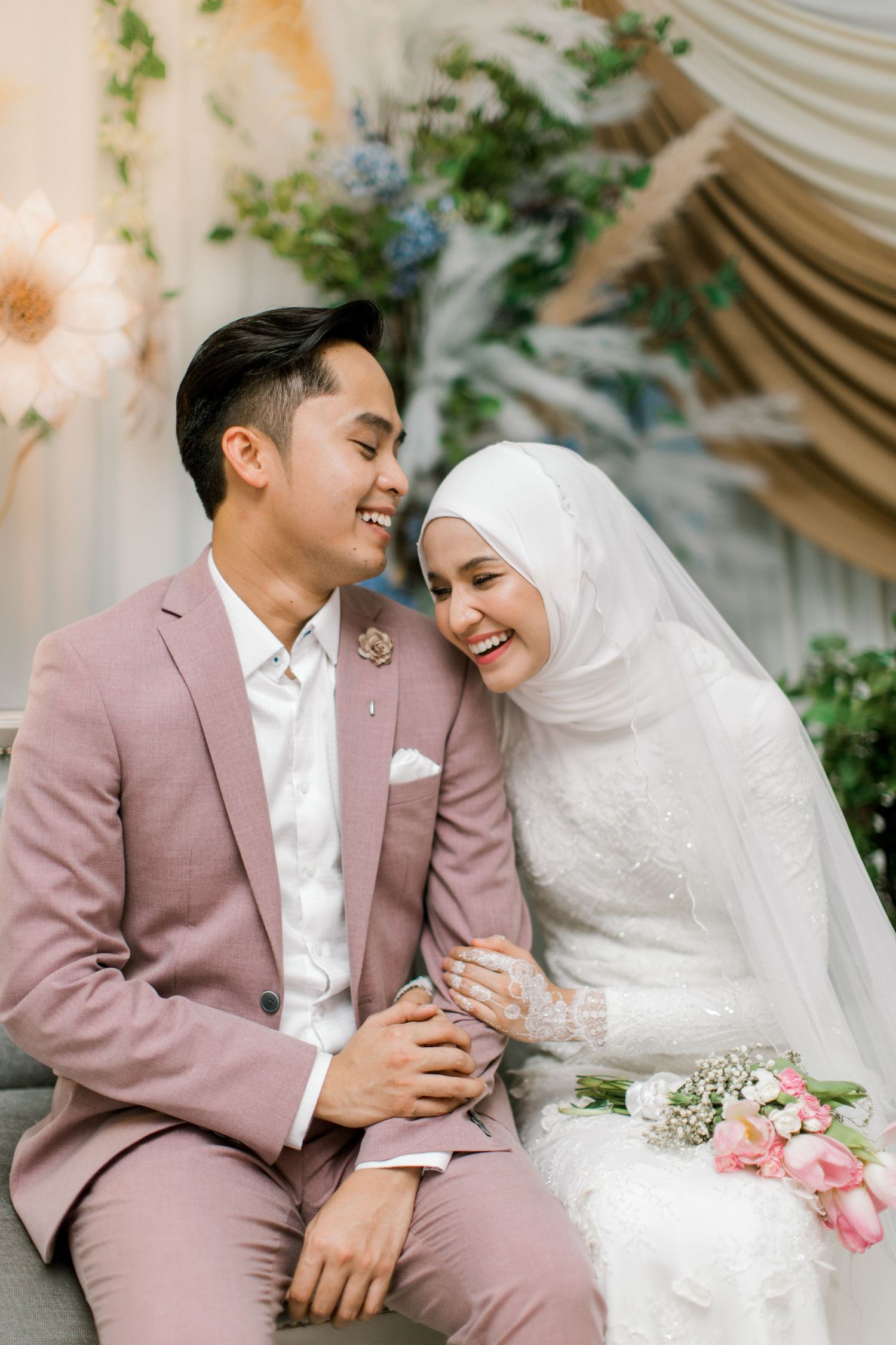 A joyful Malaysian couple in wedding attire, groom in pink suit and bride in white hijab, sharing a happy moment together.