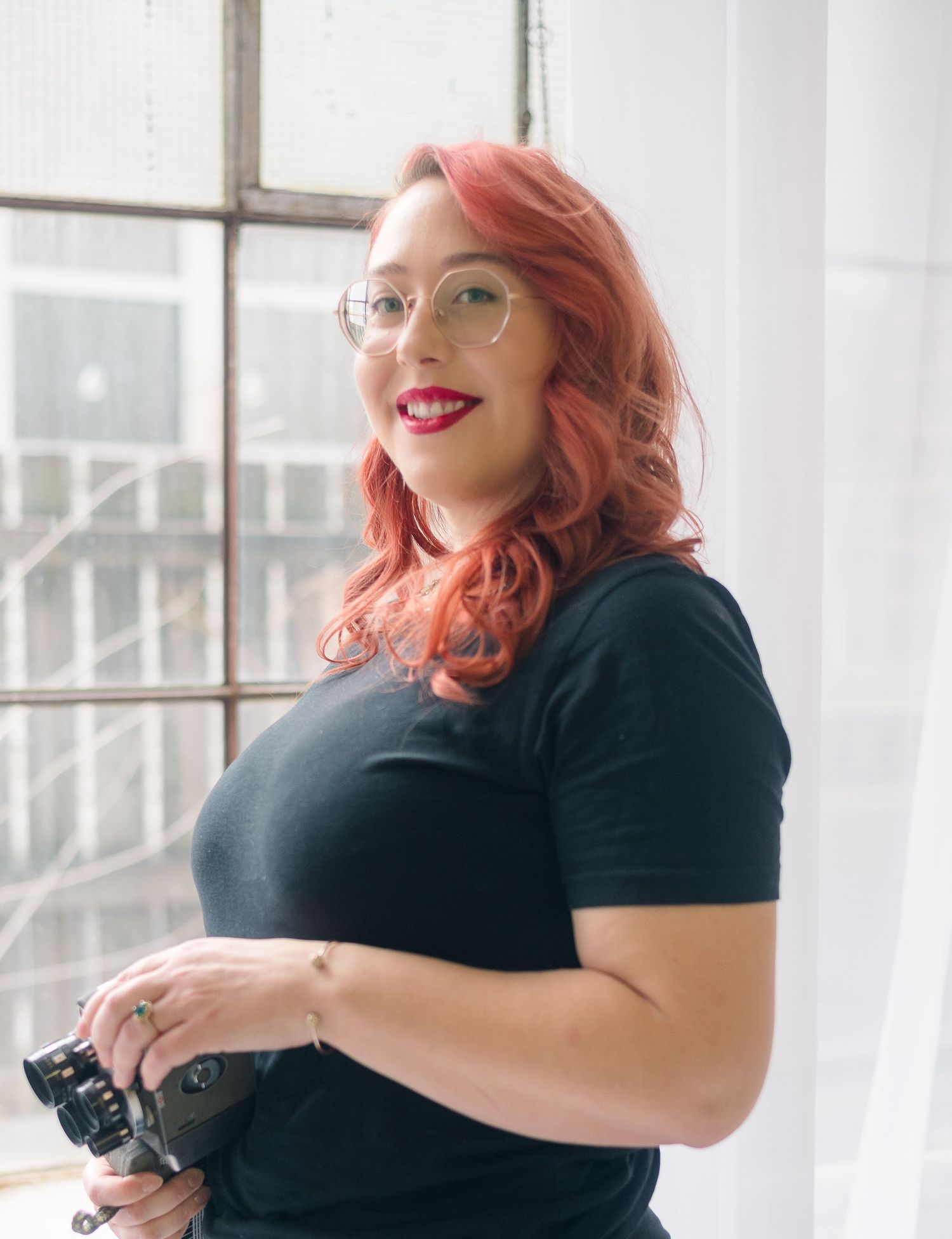 Woman with red hair and glasses holding a camera, wearing a black t-shirt, standing near a window.