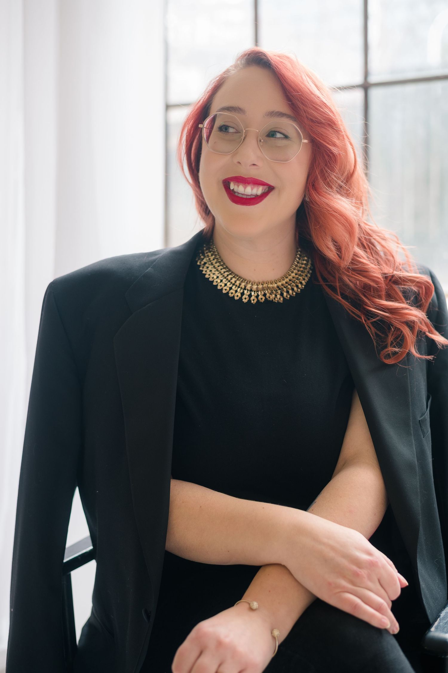 Smiling woman with long wavy red hair, glasses and red lipstick wears a black blazer and gold collar necklace beside a bright window.