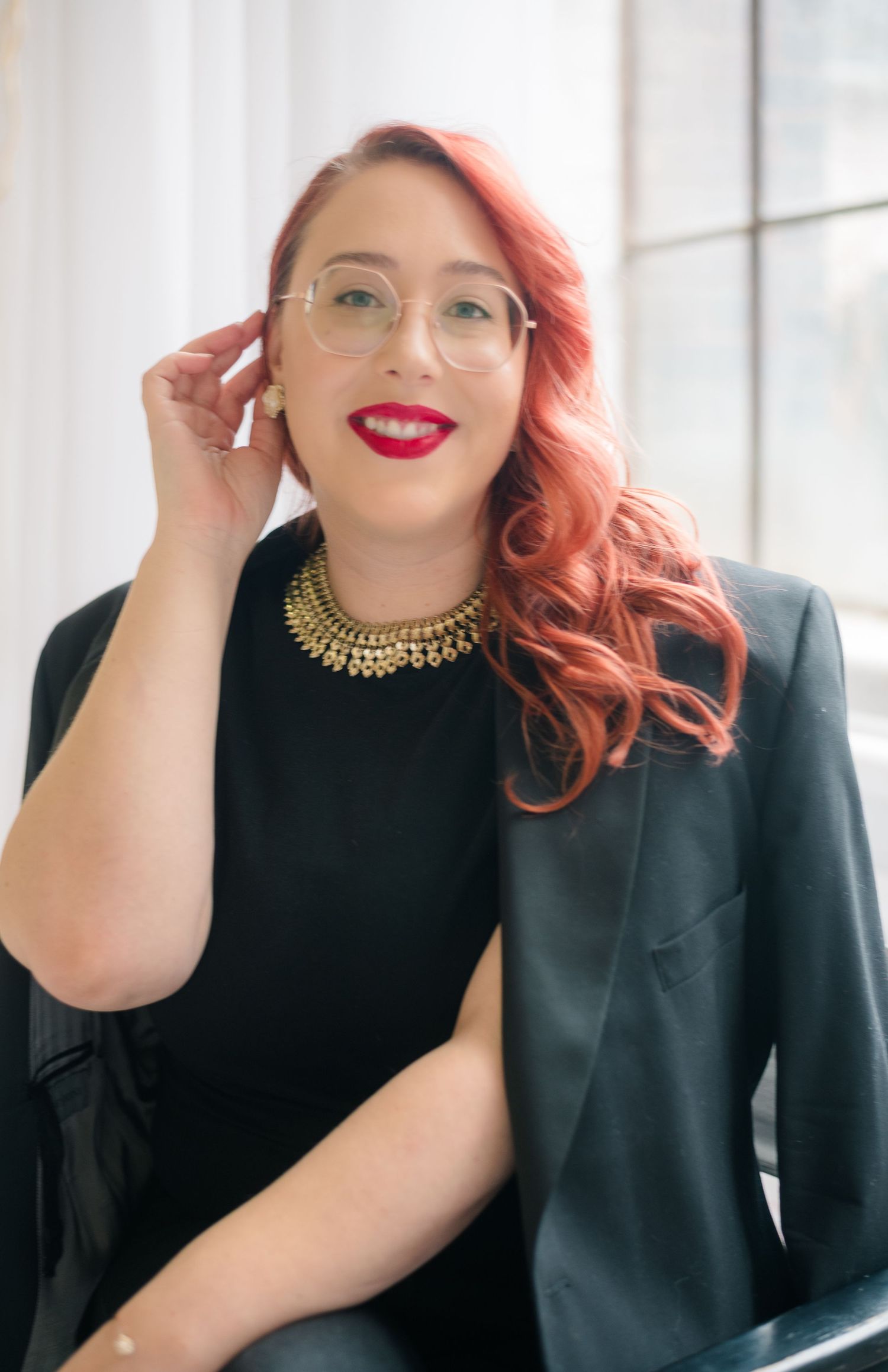 Smiling woman with wavy red hair, round glasses and red lipstick wears a gold collar necklace and black blazer while touching her ear.