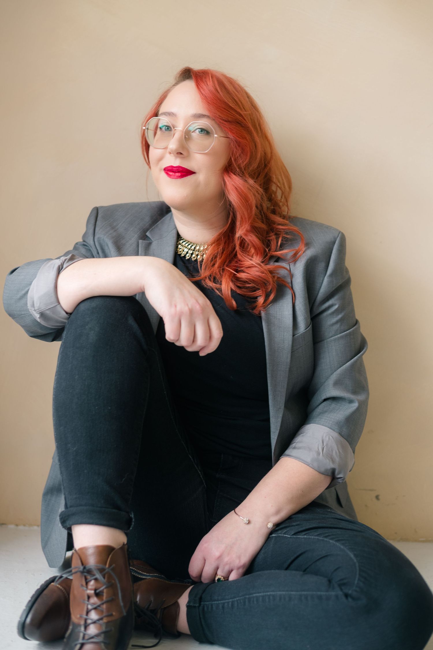 Woman with red hair and glasses sits on floor in gray blazer, black jeans, and brown boots against beige wall.