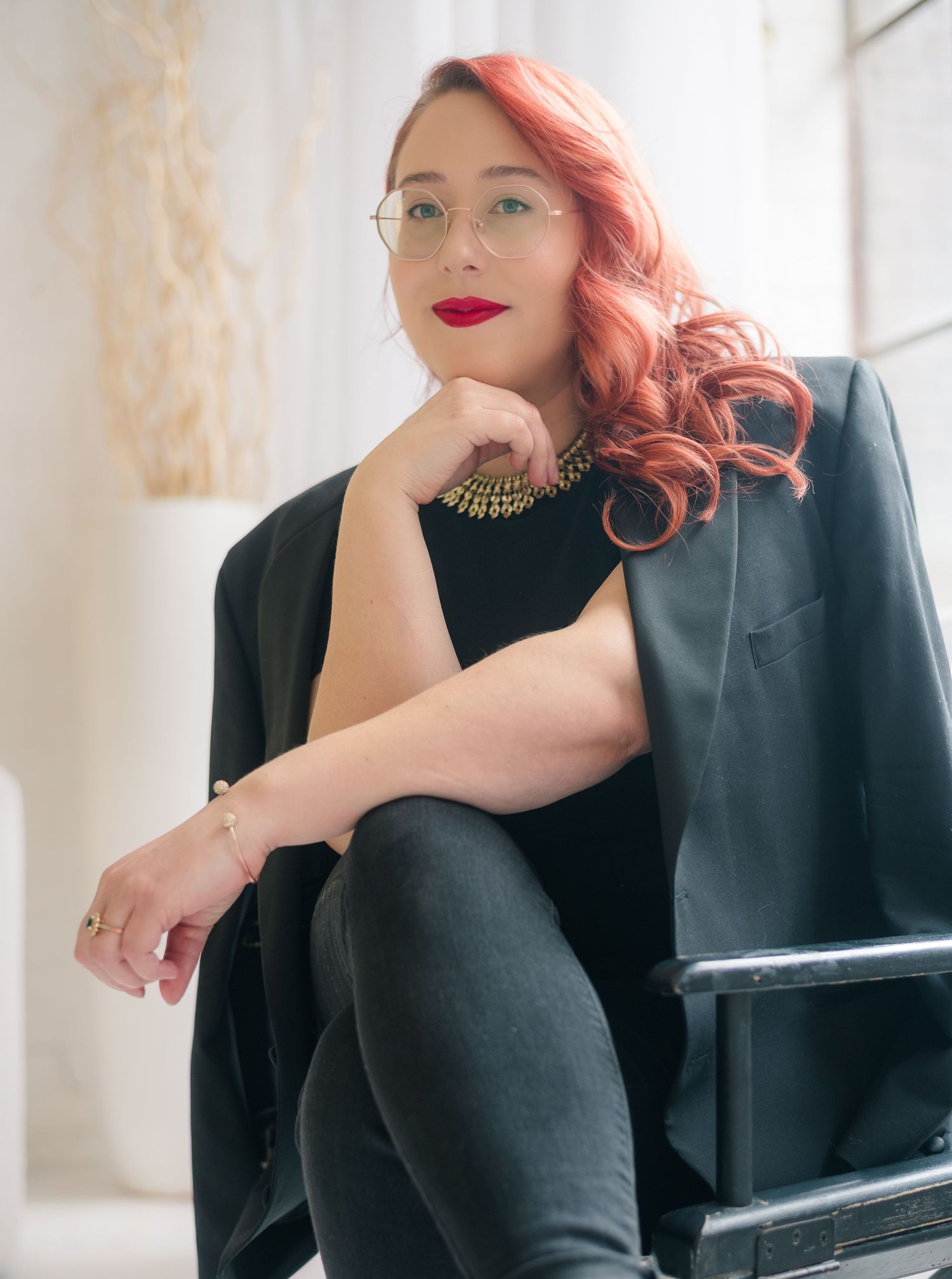 Woman with red hair and glasses wearing black blazer and gold necklace poses confidently in a chair.
