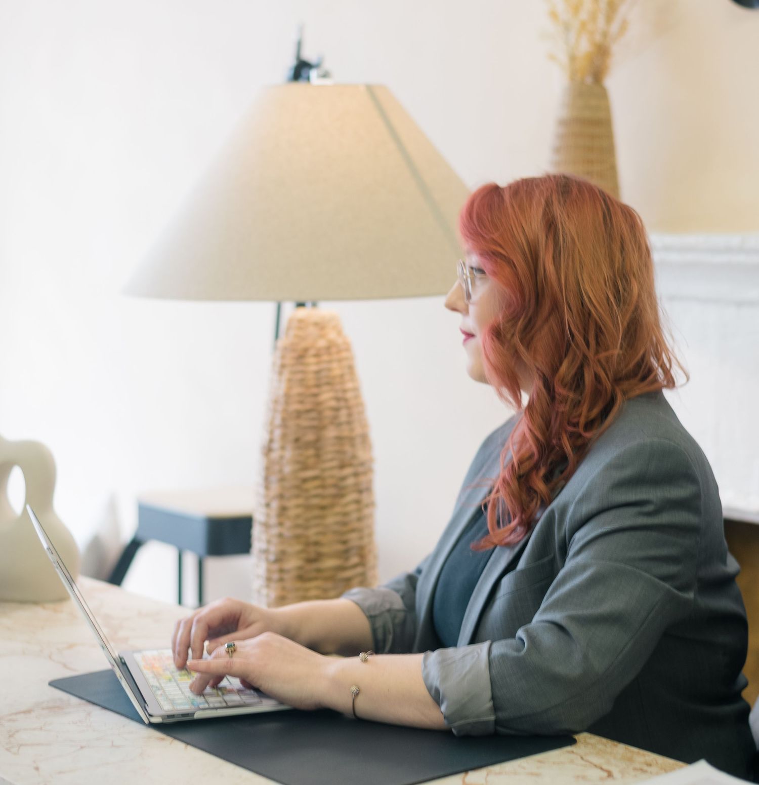 A professional woman with red hair types on a laptop at a desk, wearing a gray blazer with a lamp in the background.