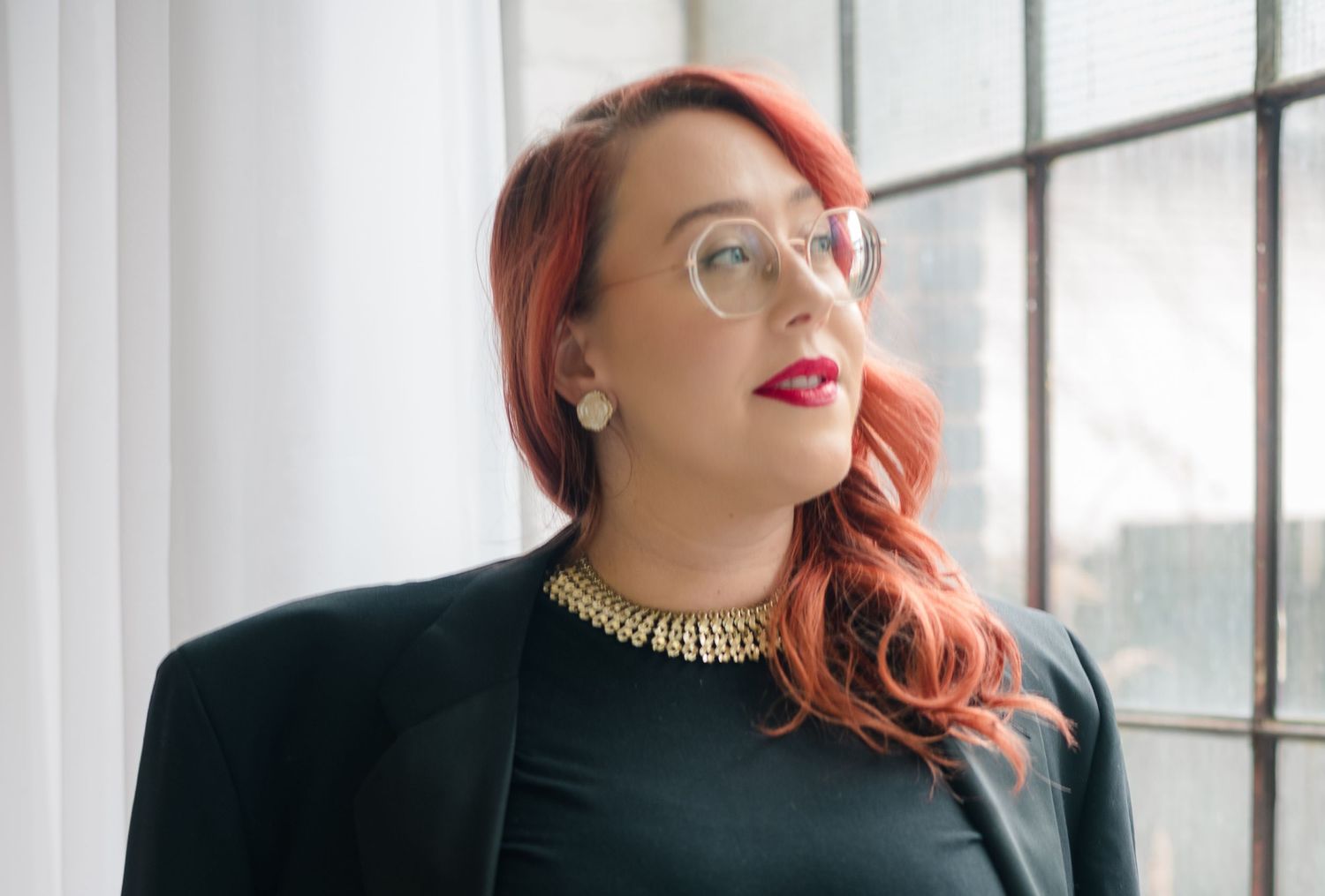 Woman with wavy auburn hair, clear glasses and red lipstick gazes toward a bright window in a black blazer and gold collar necklace.