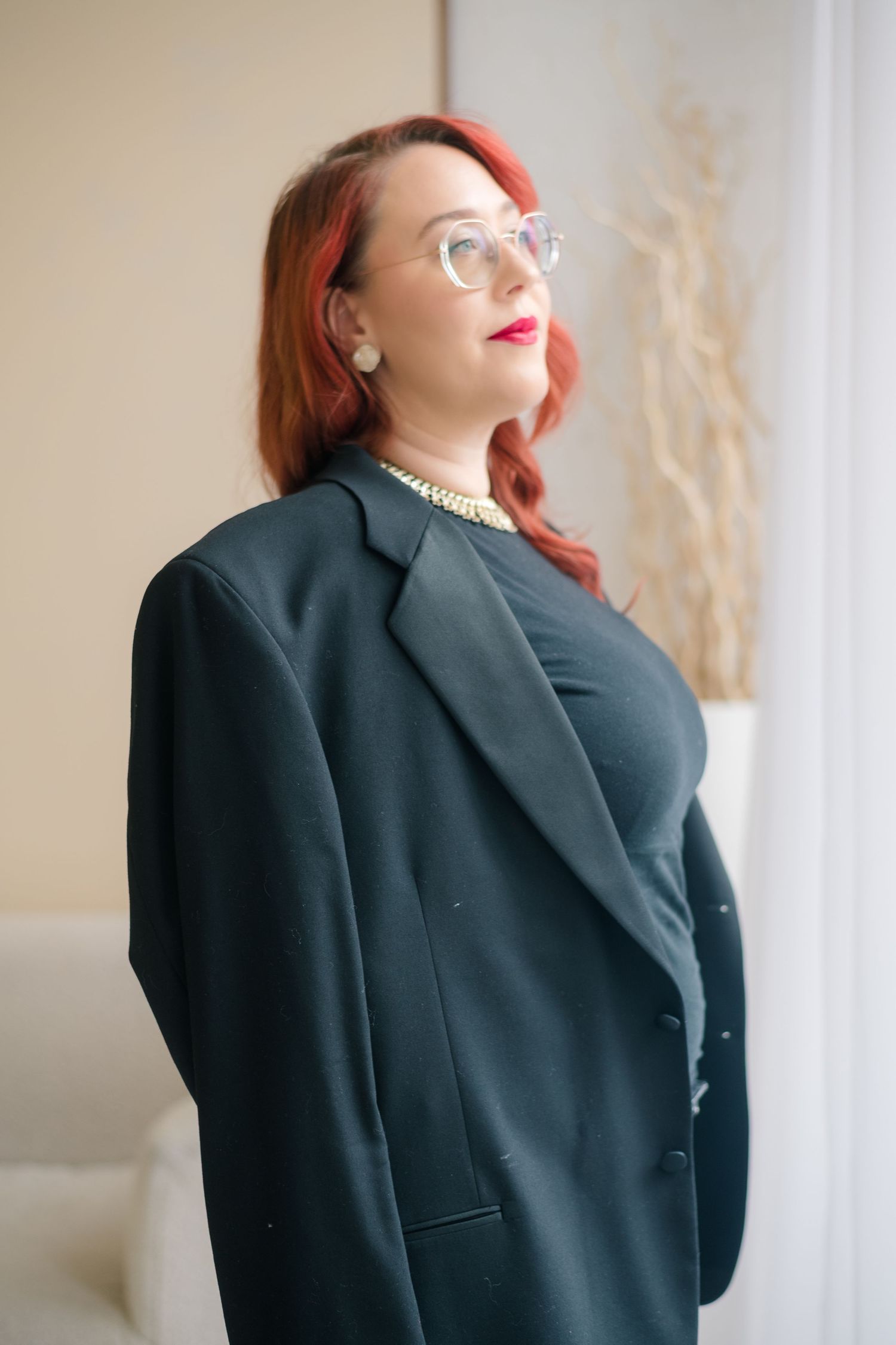 Woman with wavy red hair, round glasses, and red lipstick wears a black blazer and pearl necklace while gazing upward by a sunlit window.