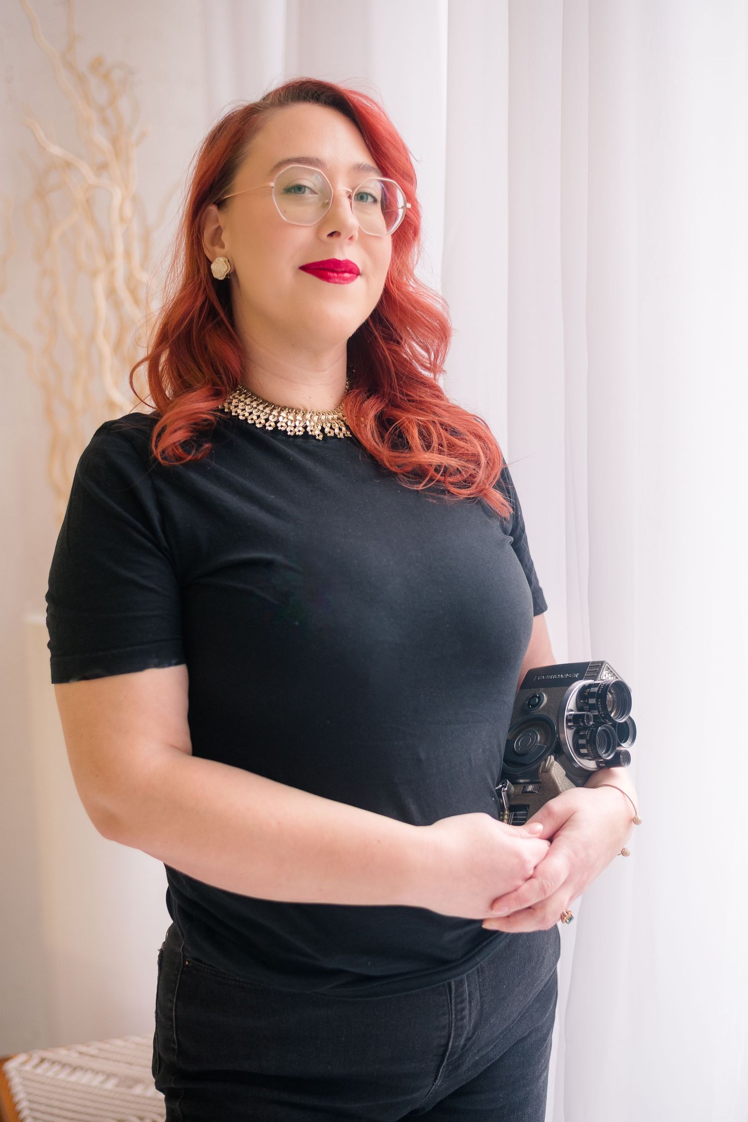Woman with red wavy hair, glasses and red lipstick wearing a black tee and pearl necklace, holding a vintage film camera by a sheer curtain.