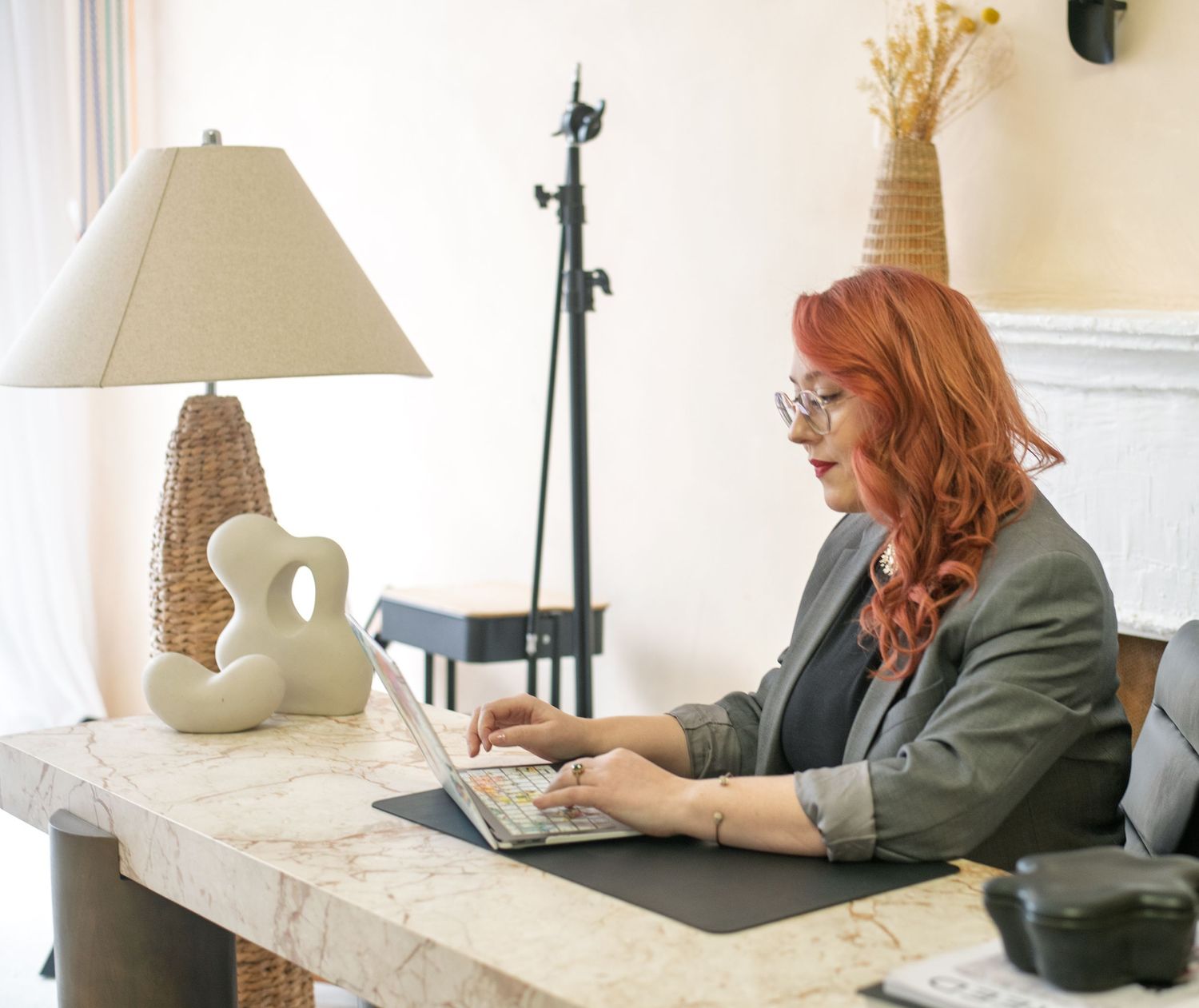 A woman with red hair wearing glasses works on a laptop at a marble desk beside a lamp in a bright home office.