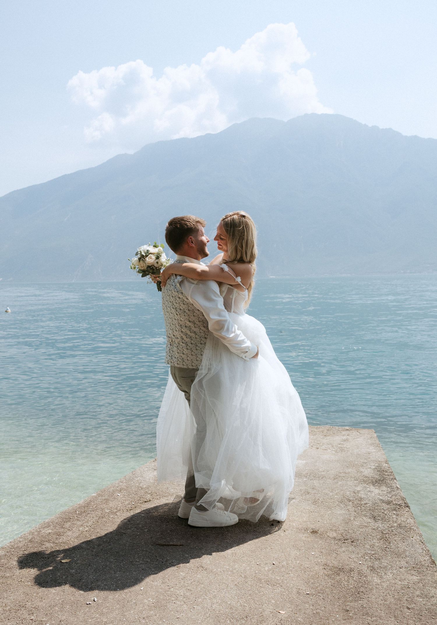 Newlywed couple embracing on a lakeside pier with mountains in the background, bride in white gown holding bouquet.