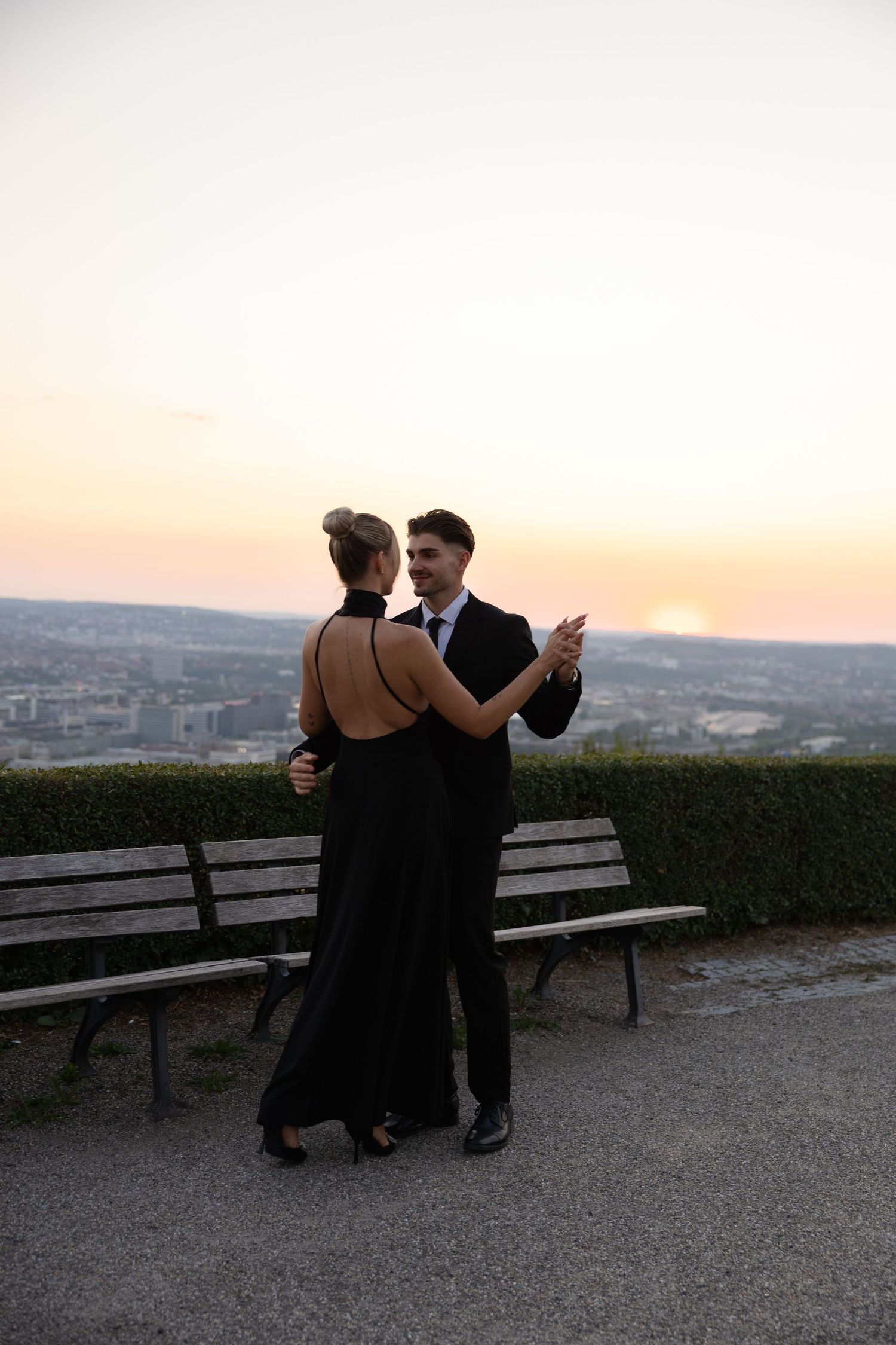 Elegant couple dancing in formal black attire at sunset with a scenic city view backdrop.