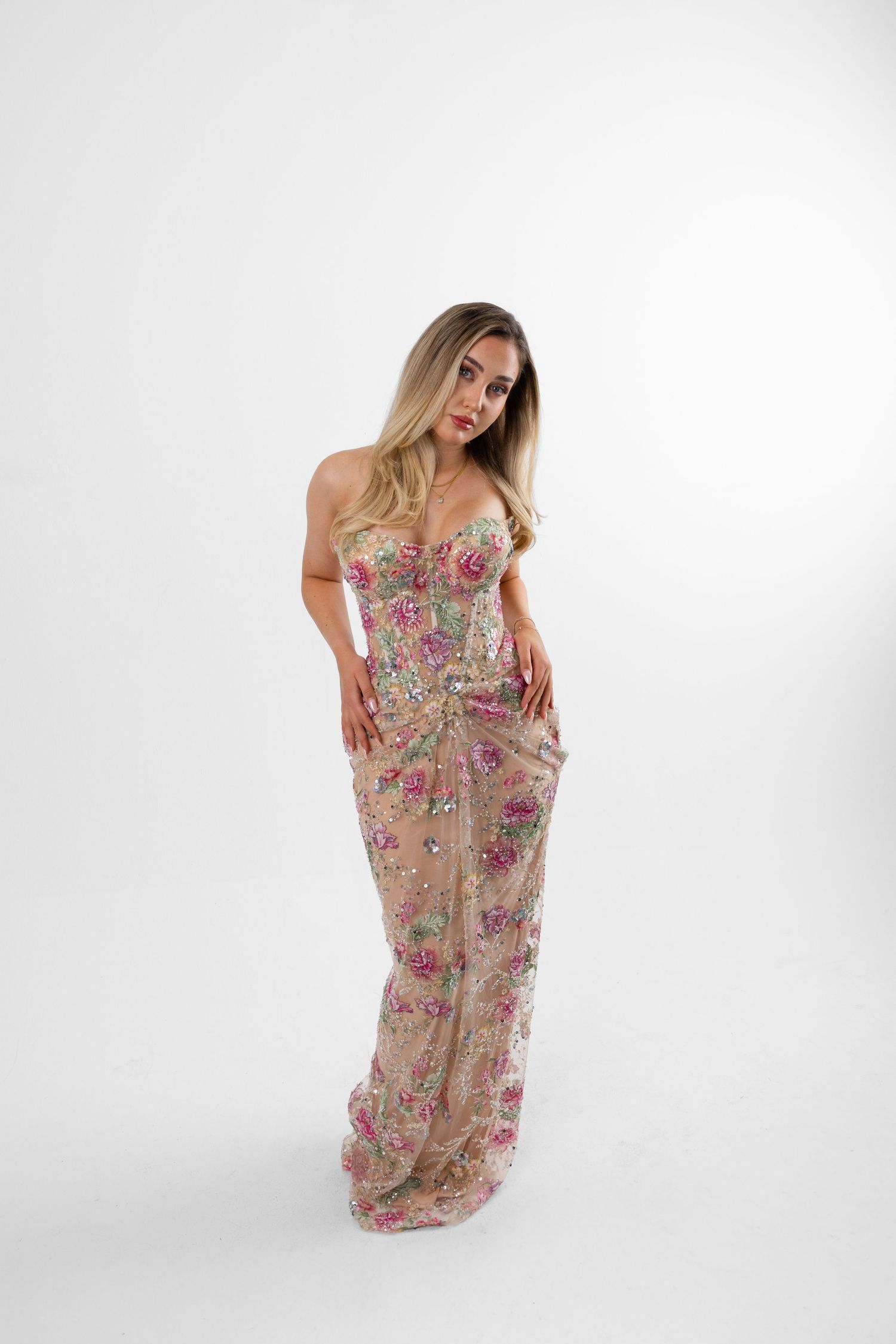 Woman in strapless floral sequin gown with pink and gold embellishments, posing against white background.