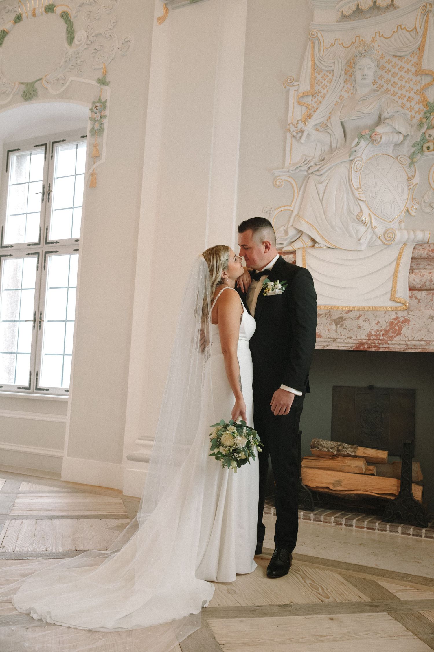 Bride and groom share a romantic moment in an elegant white room with ornate fireplace and cathedral windows.