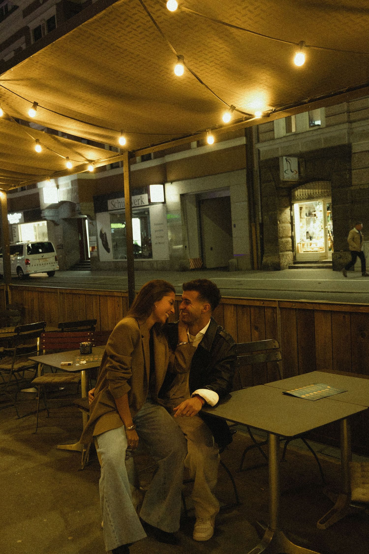 Romantic couple leaning in close at an outdoor cafe terrace under glowing string lights at night.