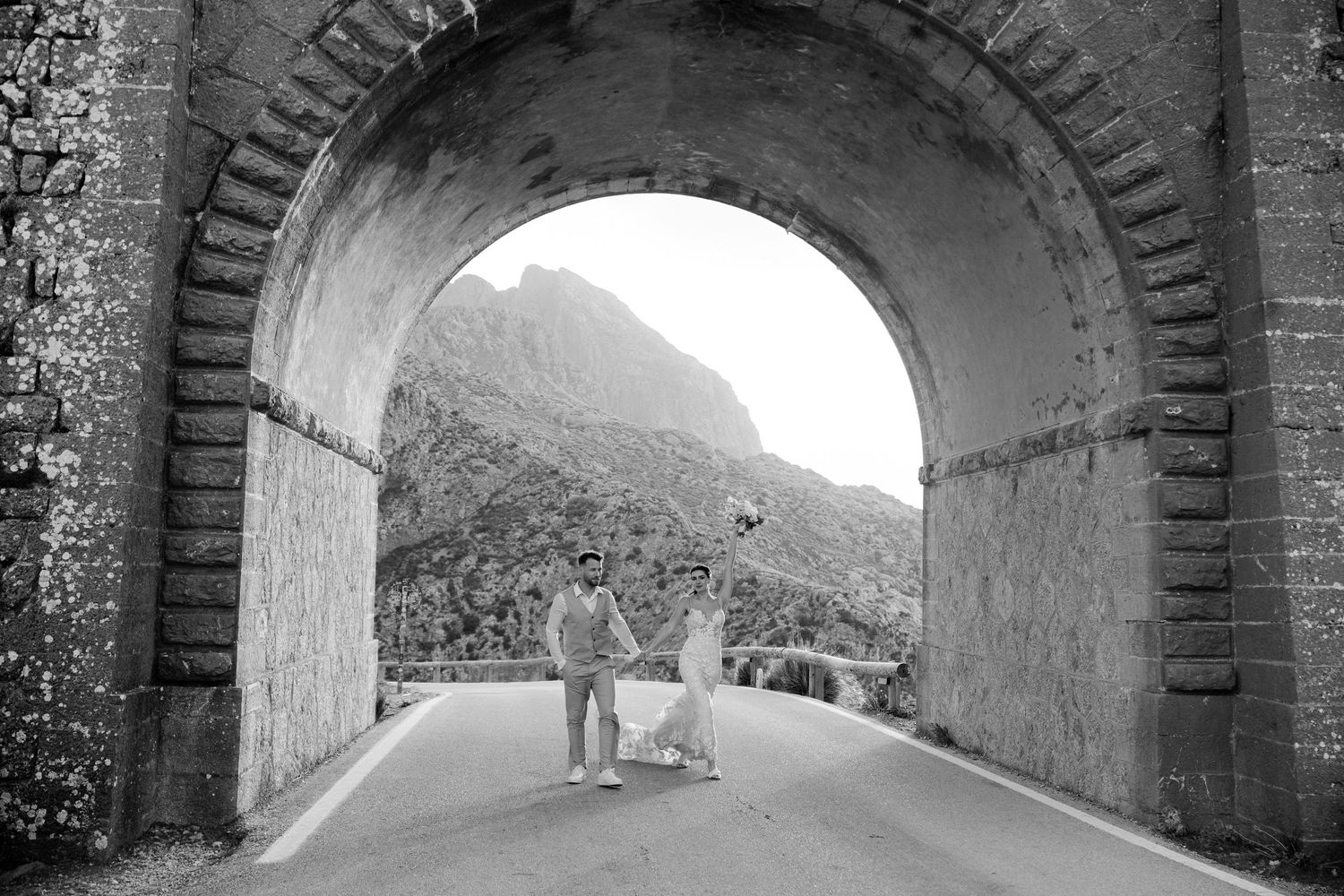 Black and white photo of groom standing alone under stone arch tunnel with mountain view beyond.