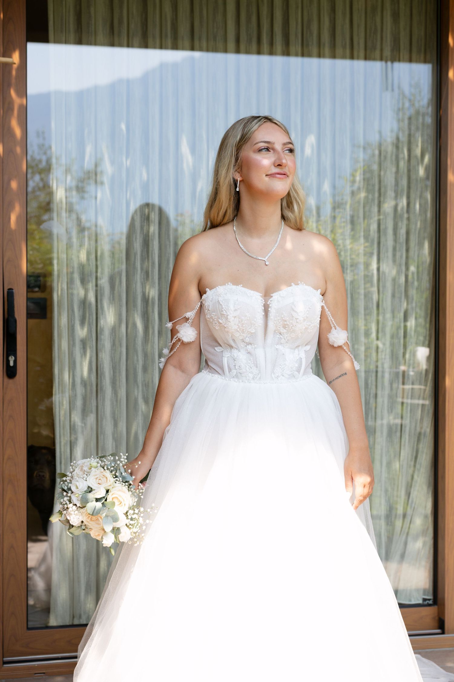 Bride in strapless lace ballgown holding white bouquet standing before glass doors with cactus reflection.