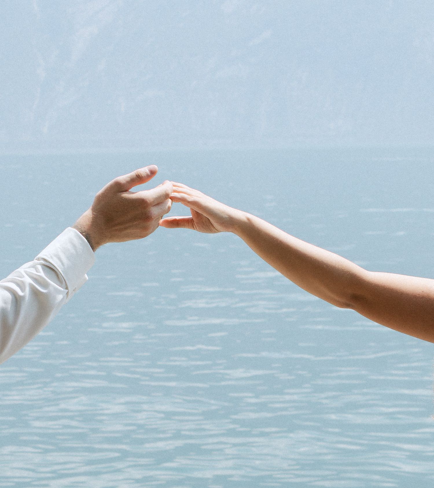 Two hands gently touching fingertips over a serene lake with misty mountains in the background.