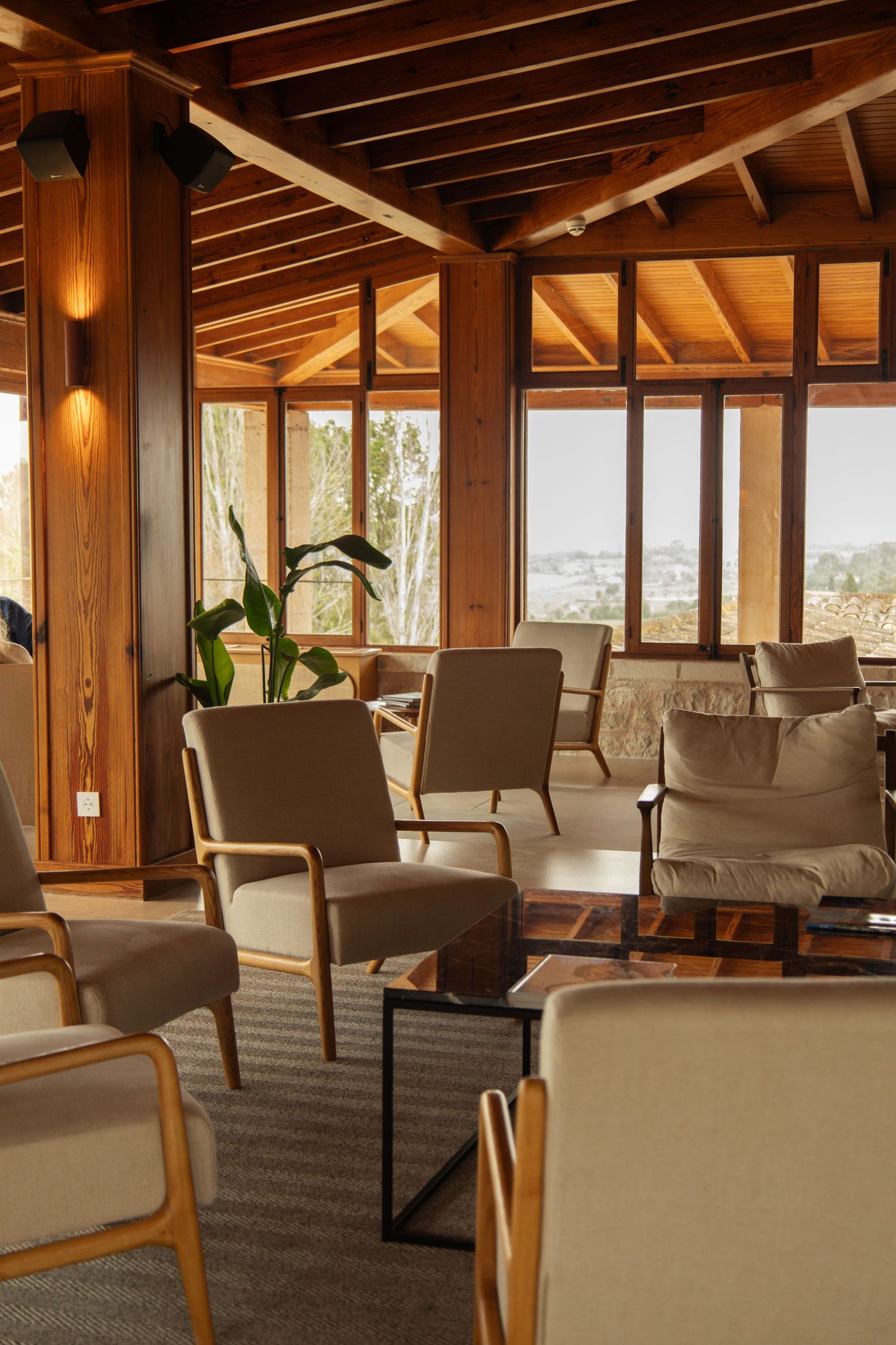 Warm wooden interior lounge with beige armchairs, exposed beam ceiling, and panoramic garden view windows.