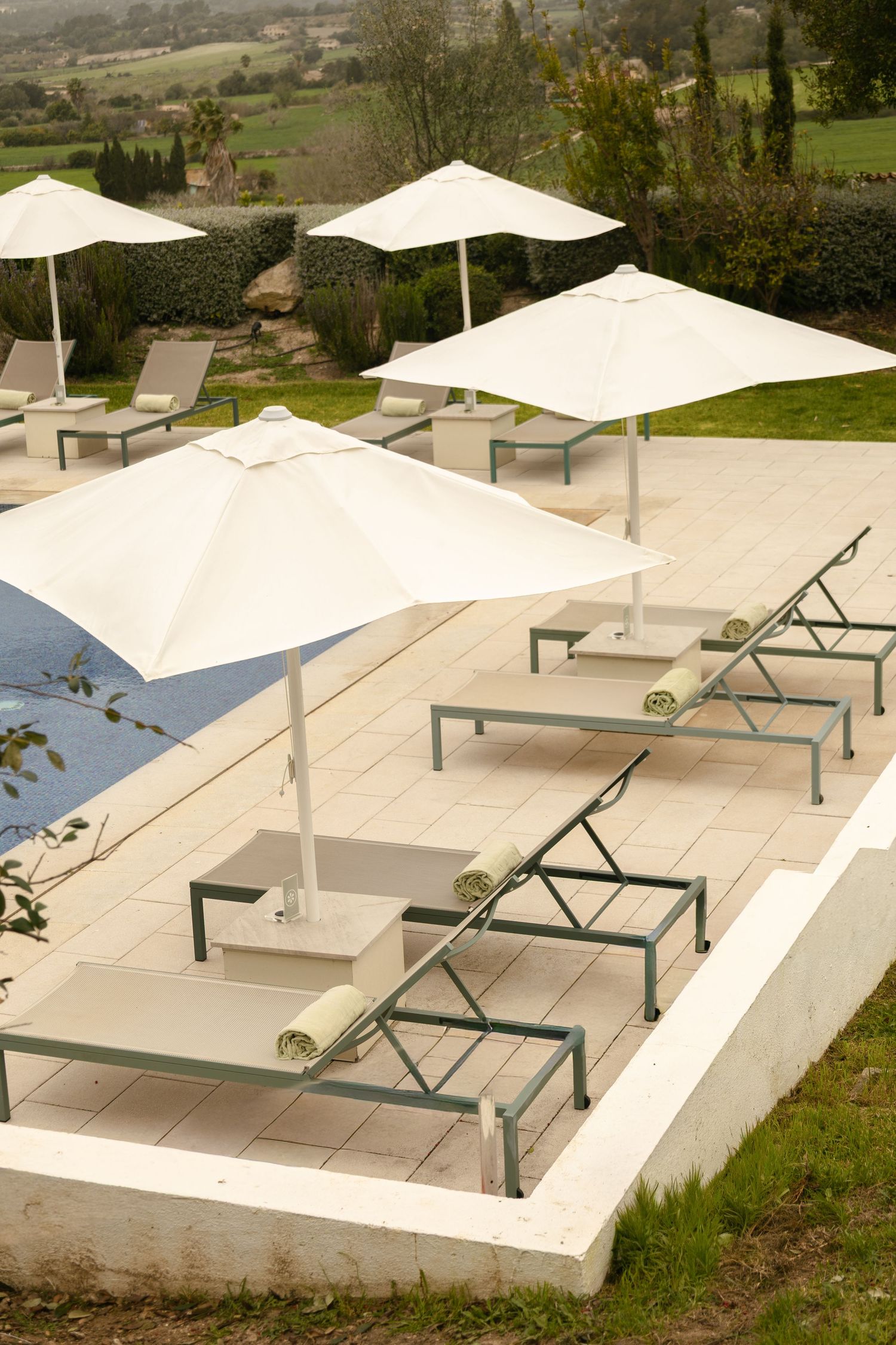 Row of white sun umbrellas and lounge chairs arranged beside a pool with green countryside views.