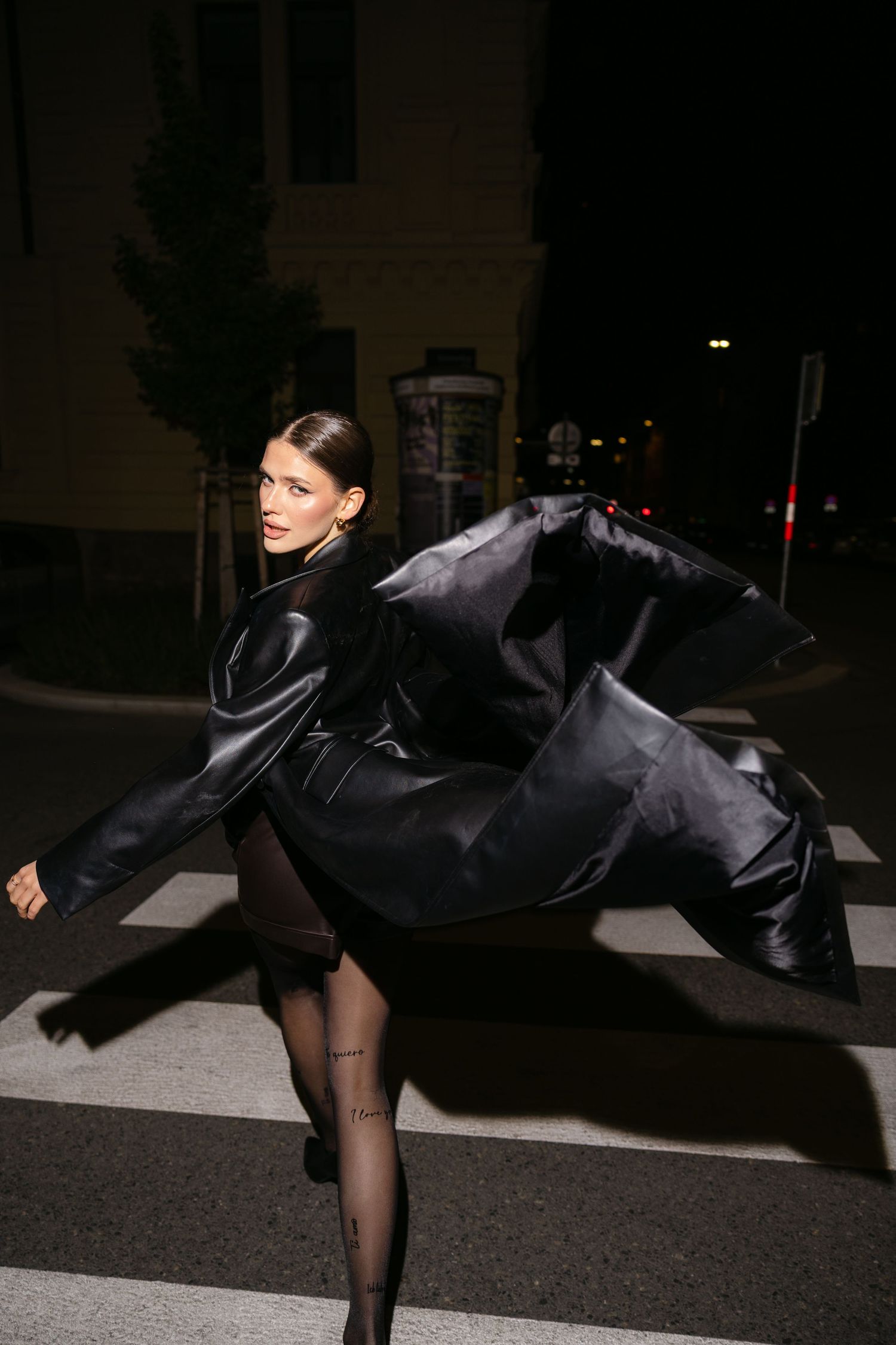 Woman in black leather coat walking across street crosswalk at night with coat flowing dramatically.