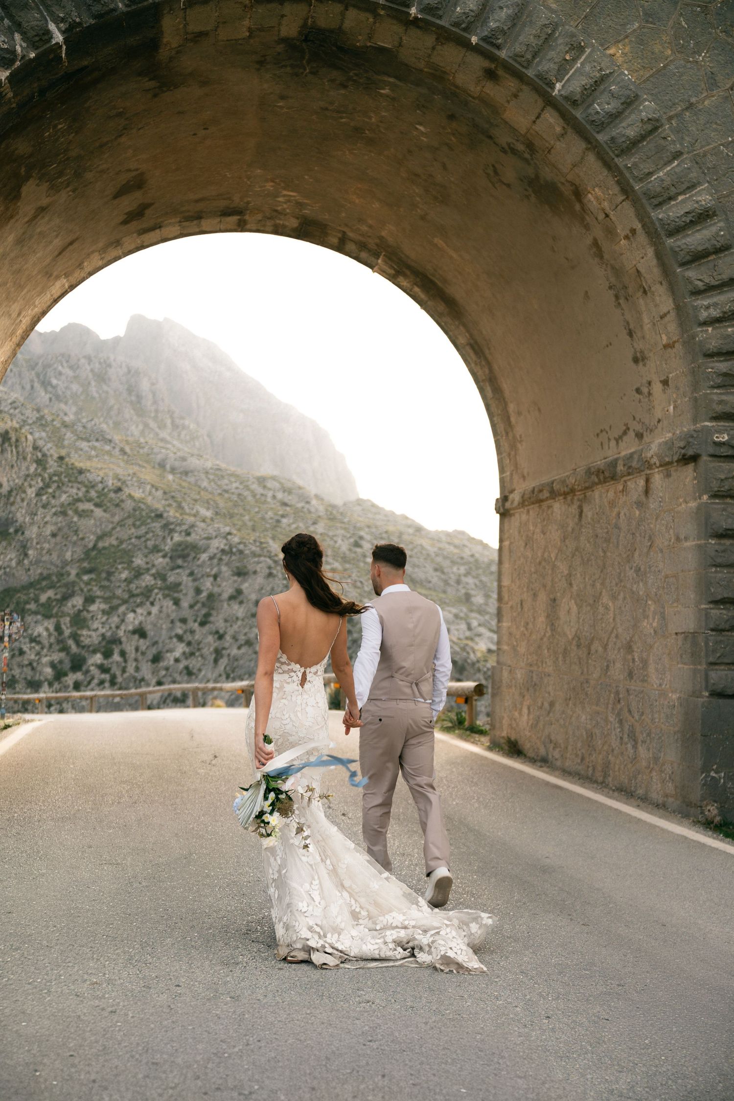 Bride and groom walking hand in hand through stone arch tunnel with mountain landscape beyond.