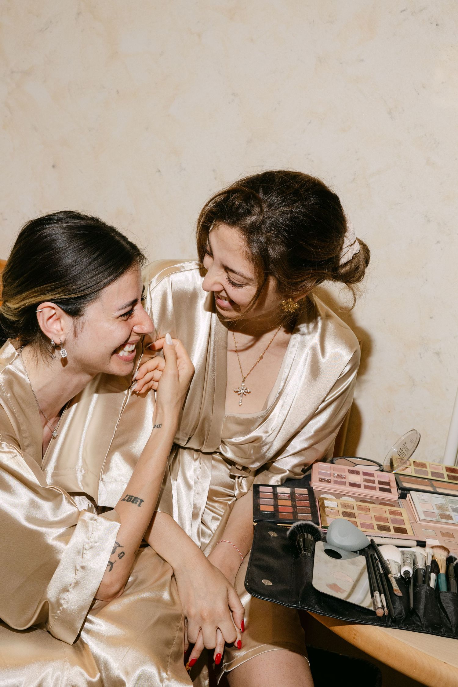 Two women in satin robes laugh together during bridal getting-ready session with makeup palettes nearby.