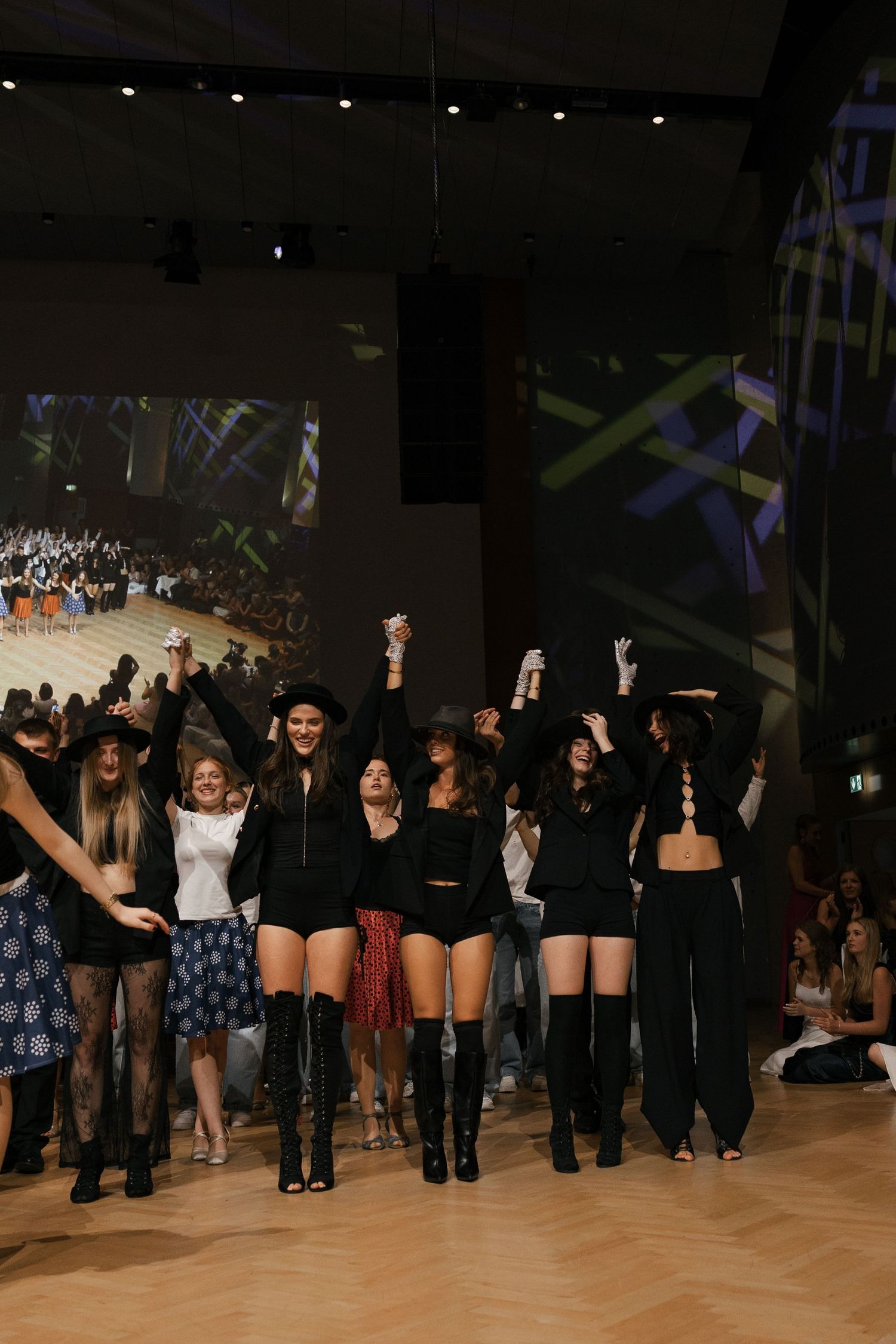 Group of performers in black outfits celebrating with raised hands on stage with projections.