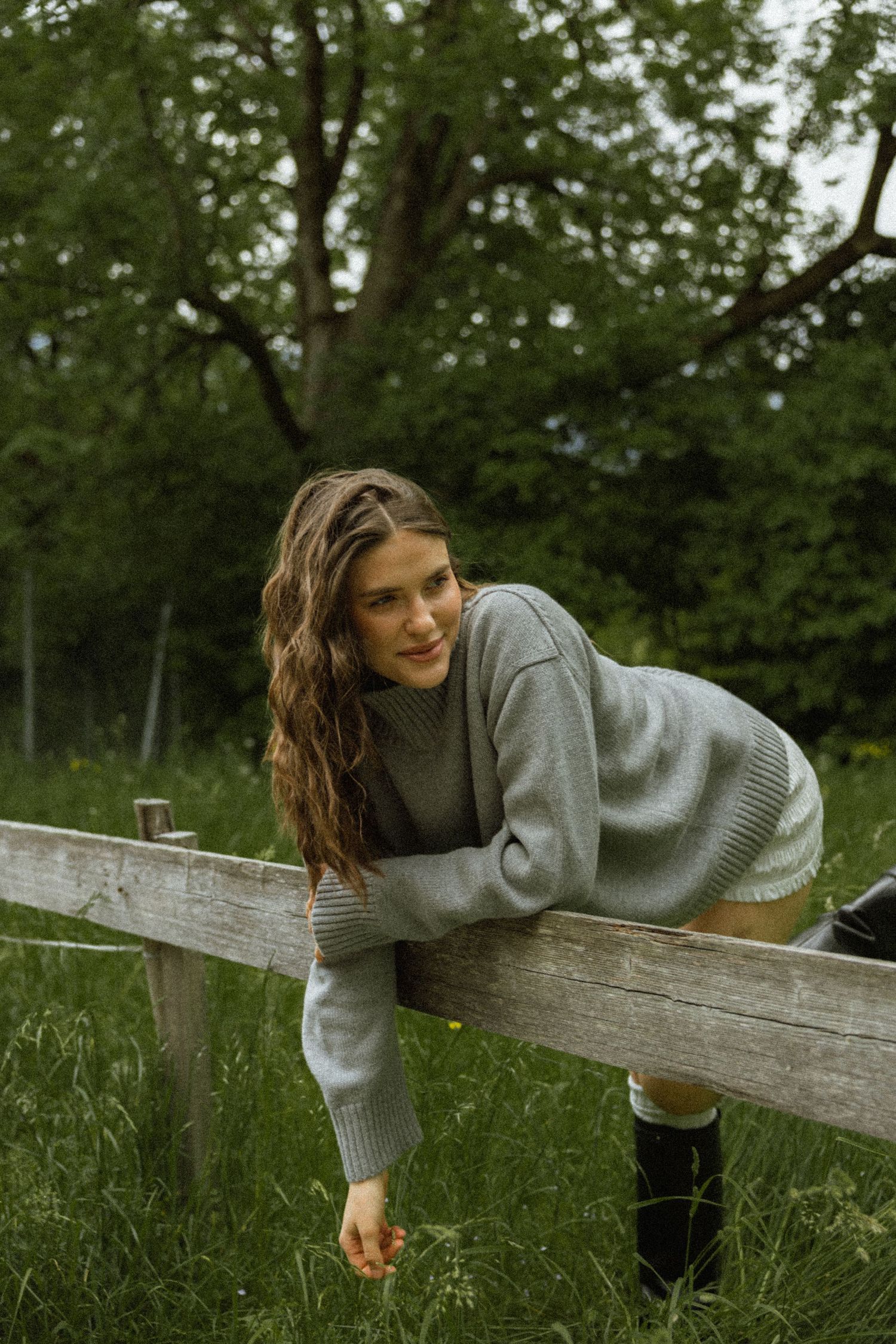 Smiling woman in grey sweater leaning over wooden fence in lush green meadow.