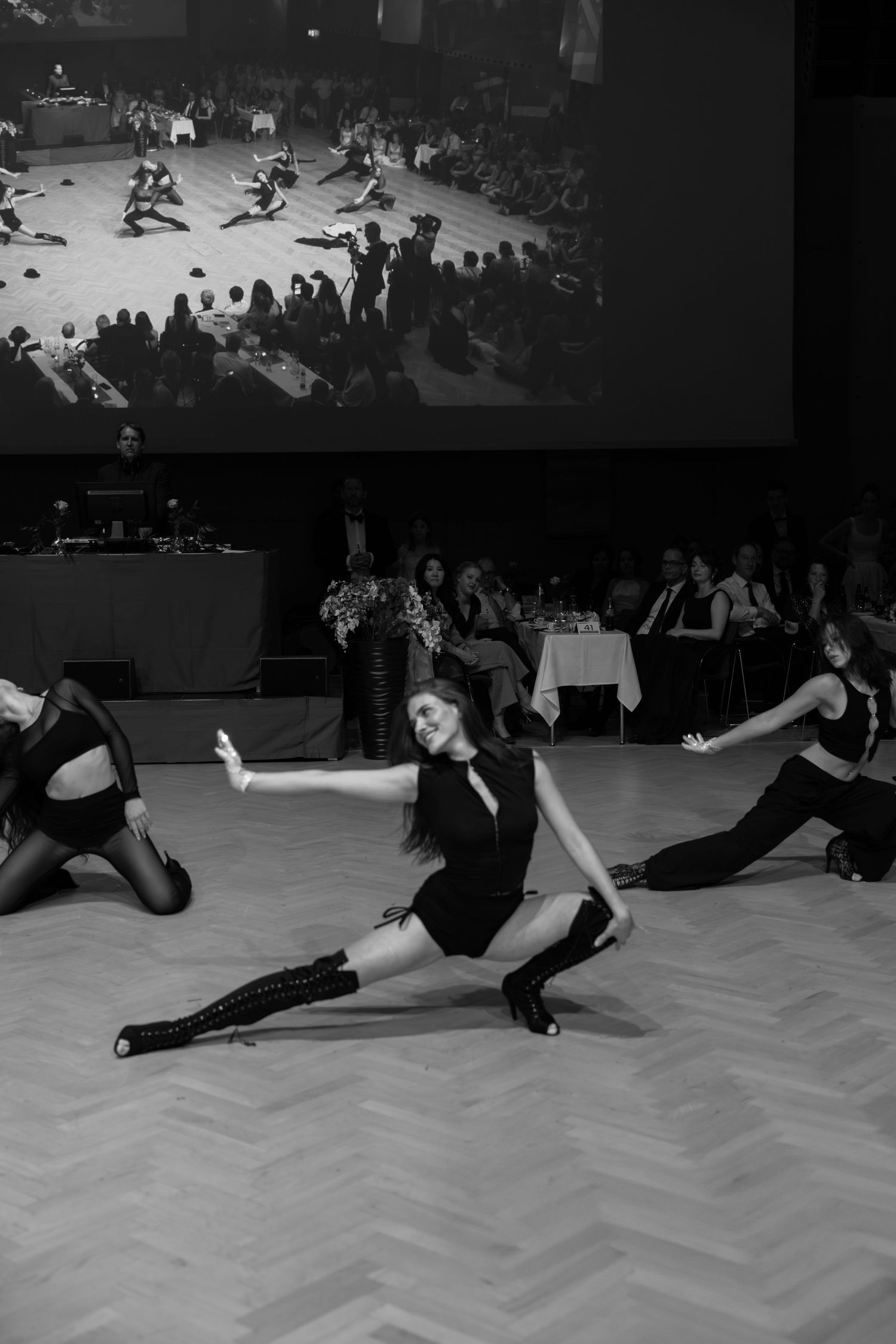Black and white photo of dancers performing an energetic routine on a ballroom floor before an audience.