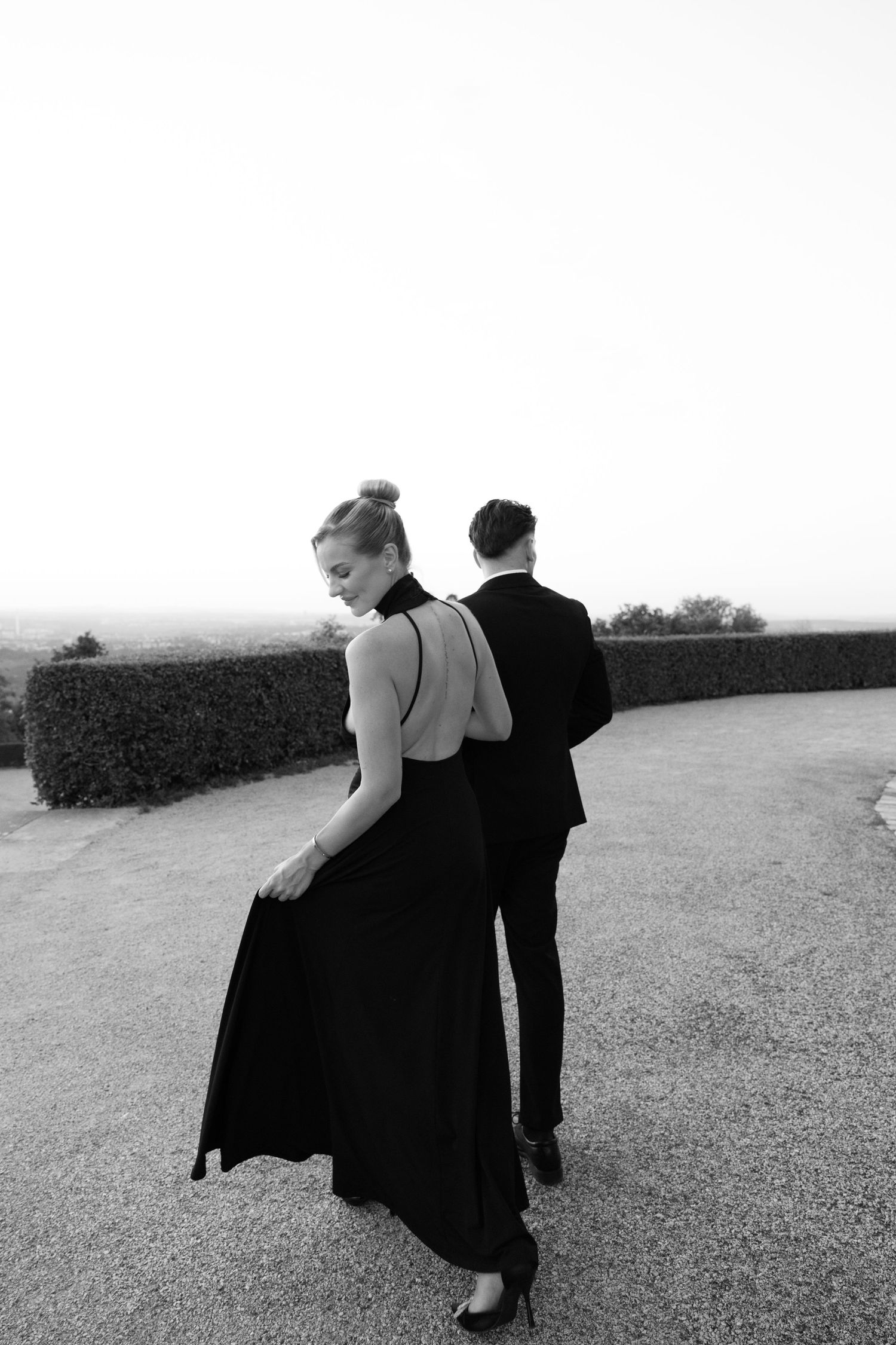 Black and white photo of an elegant couple outdoors, woman in backless black gown, man in suit, hedge backdrop.