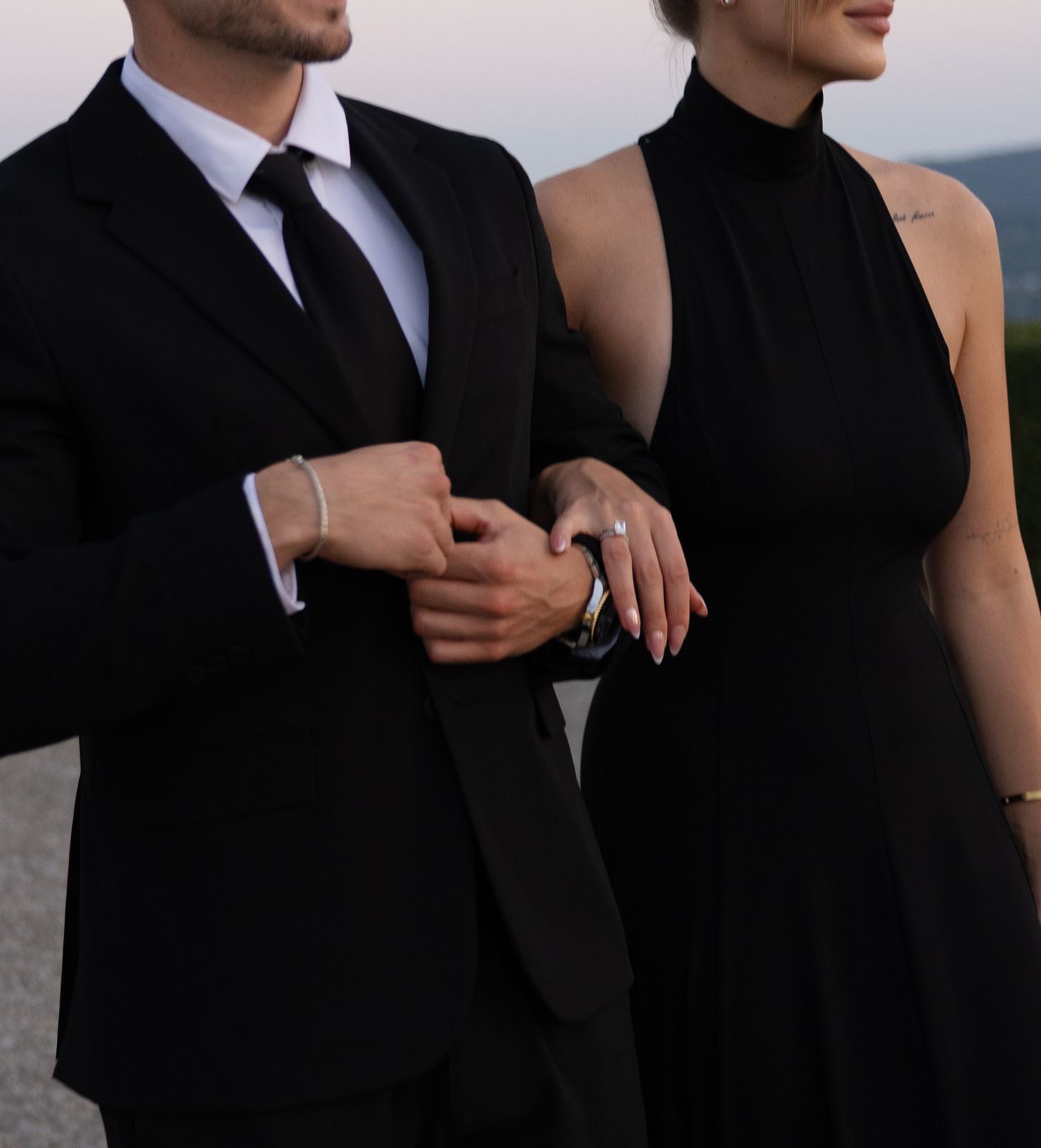 Elegant couple in matching black formal attire, man in suit with tie, woman in halter dress, posing together outdoors.