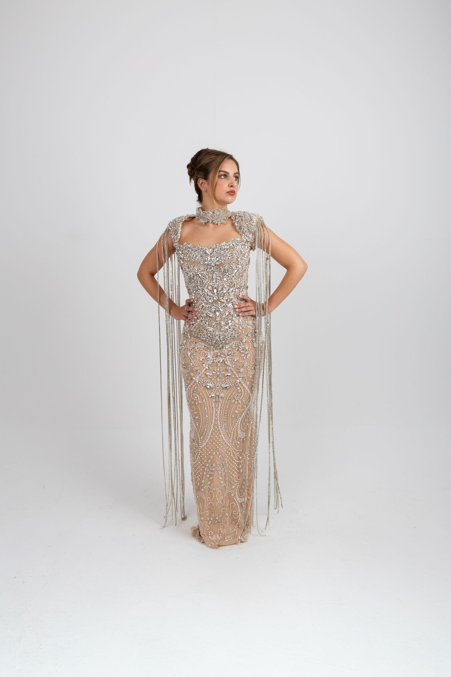 Woman in beige crystal-embellished gown with fringe cape and matching jewelry, posing on white background.
