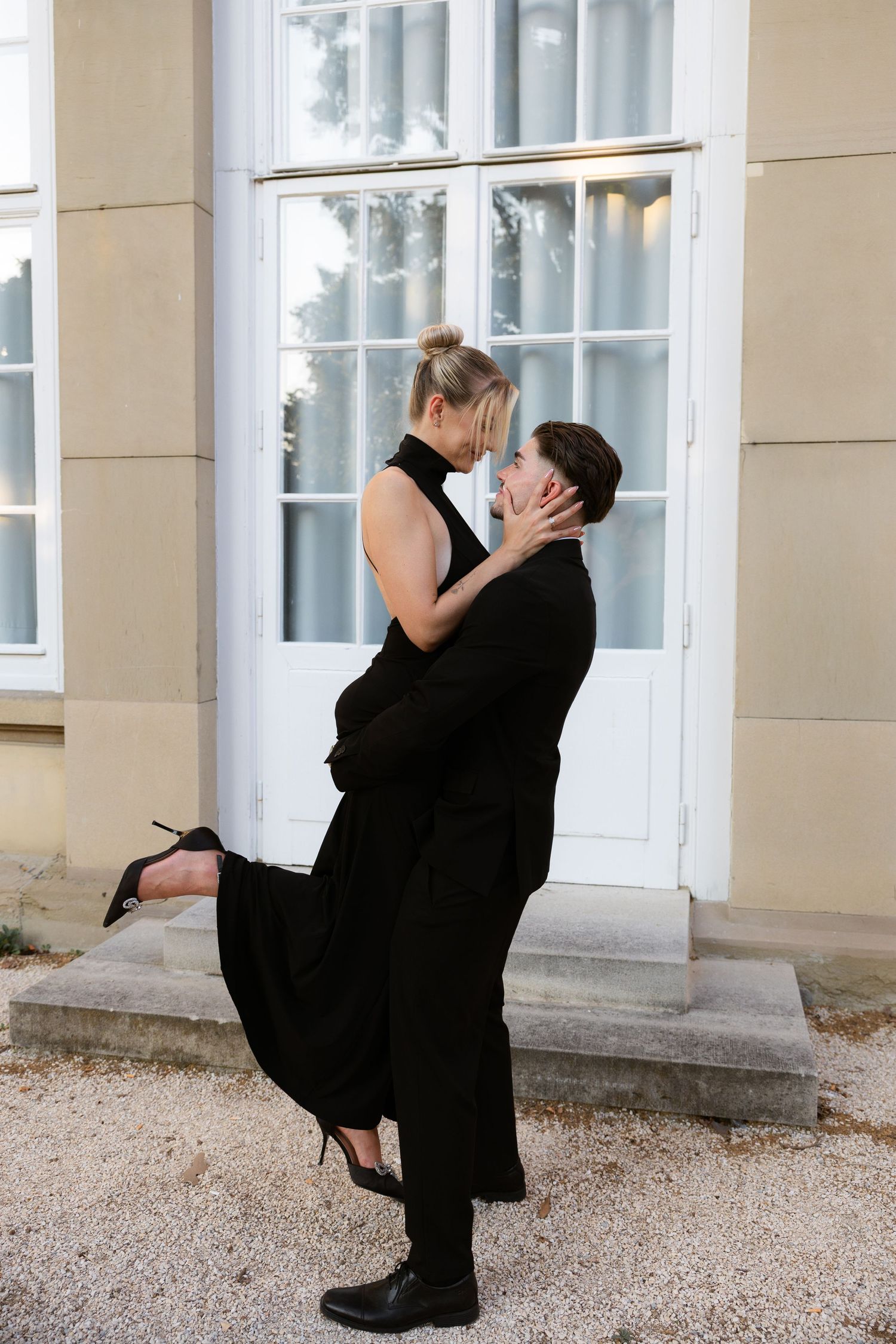 Man in black suit lifts woman in black halter dress as they lean in to kiss by a grand building.