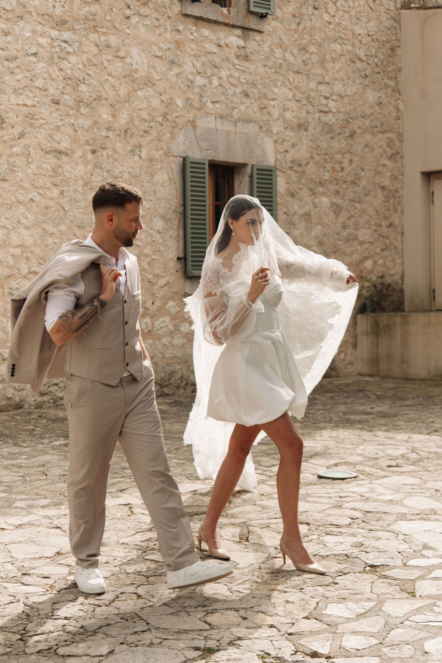 Groom in beige suit walks beside bride in short white dress with veil flowing.