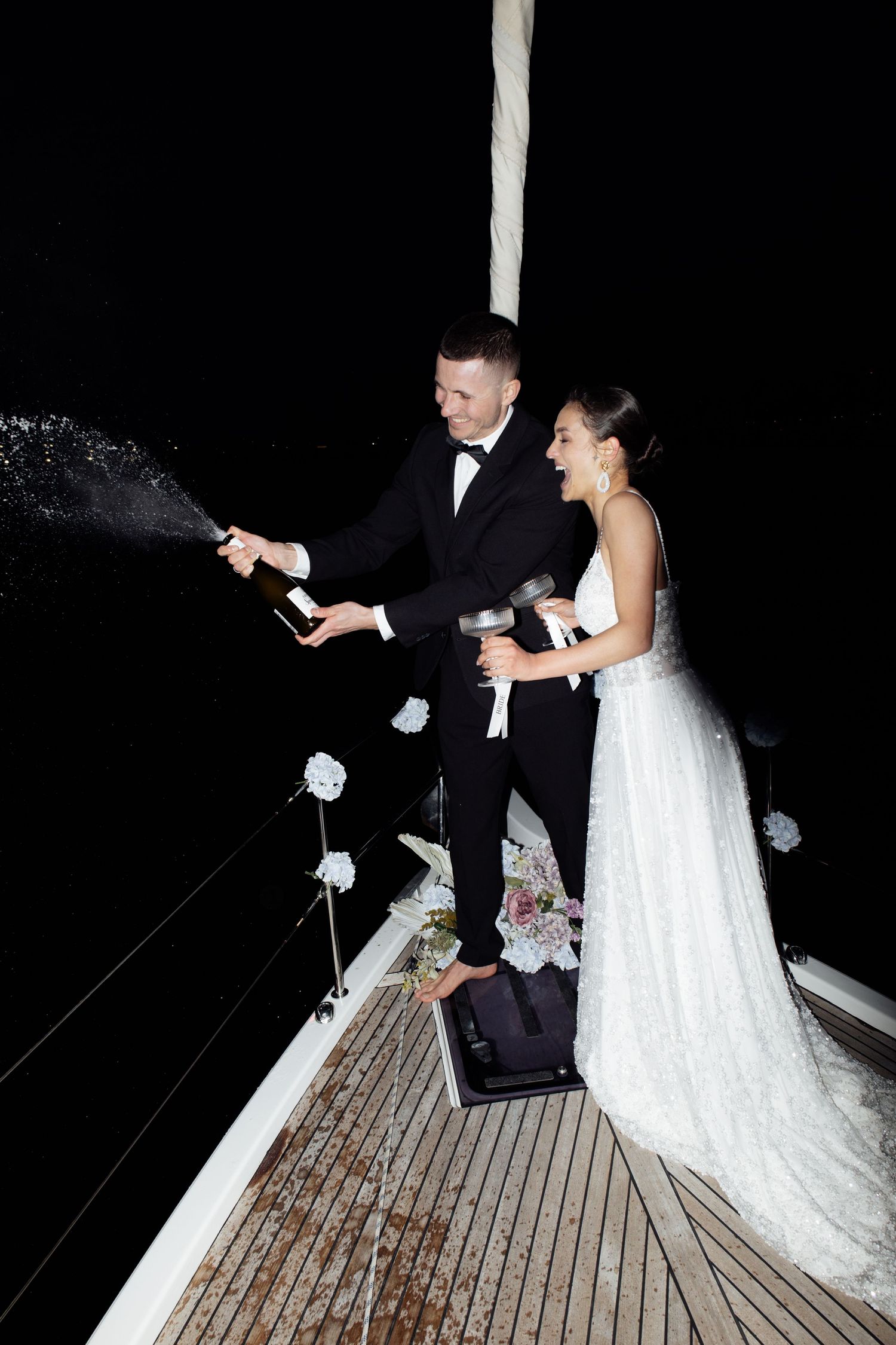 Bride and groom popping champagne on a yacht deck at night, celebrating their wedding in elegant attire.