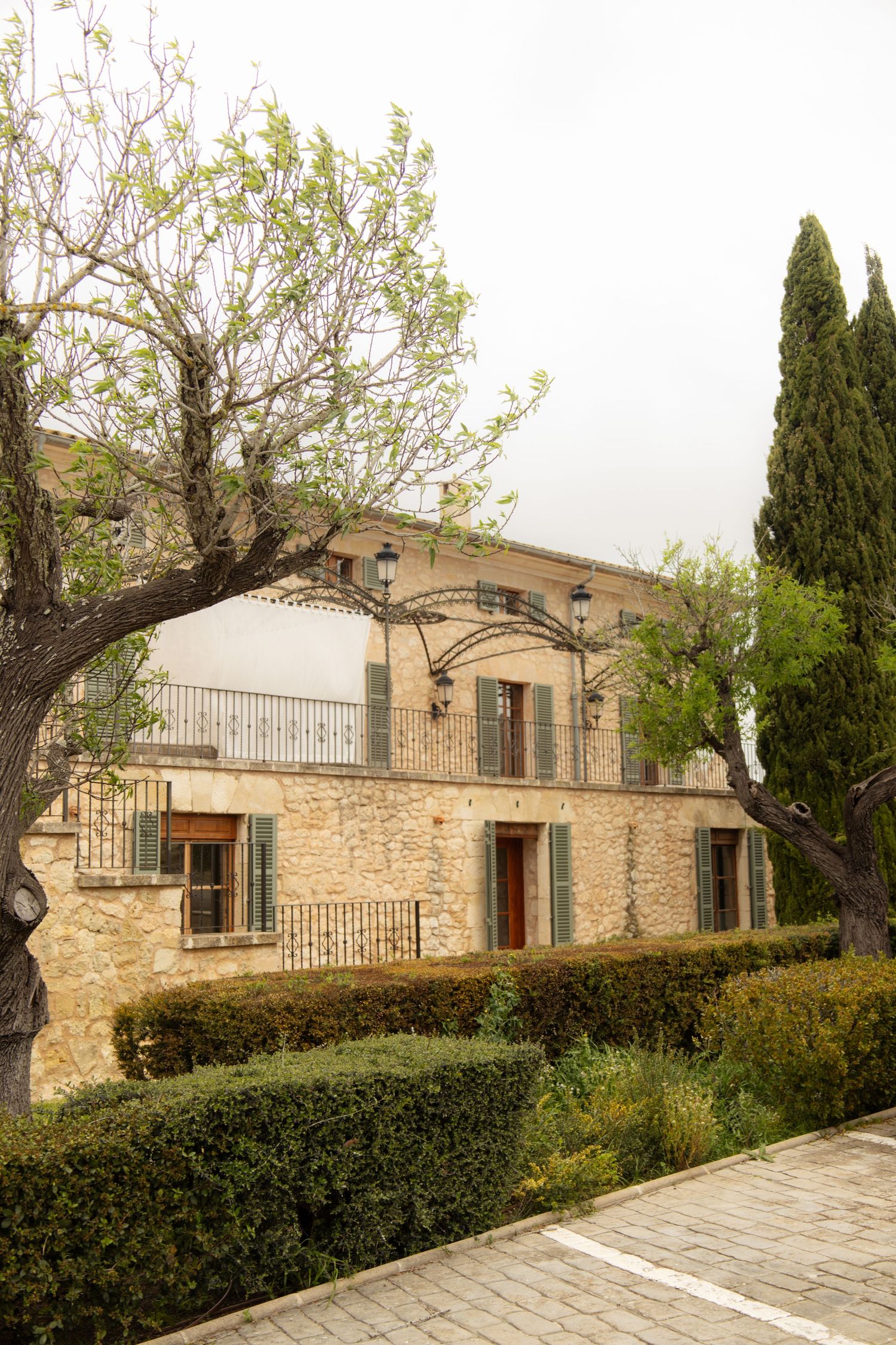 Rustic stone villa with green shutters, balcony, trimmed hedges, and cypress trees on overcast day.