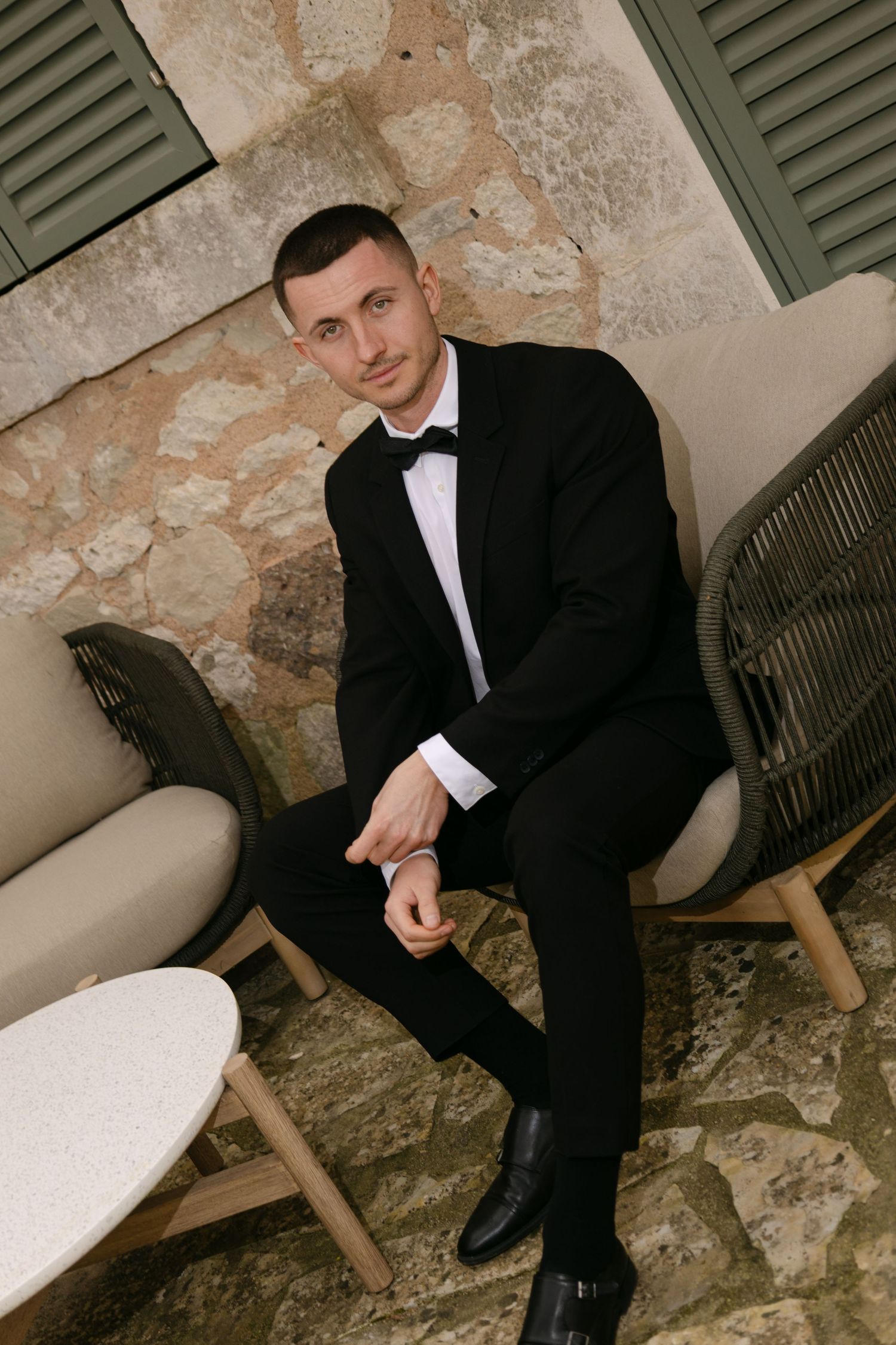 A man in a black tuxedo with bow tie sits confidently in an outdoor chair against a stone wall.