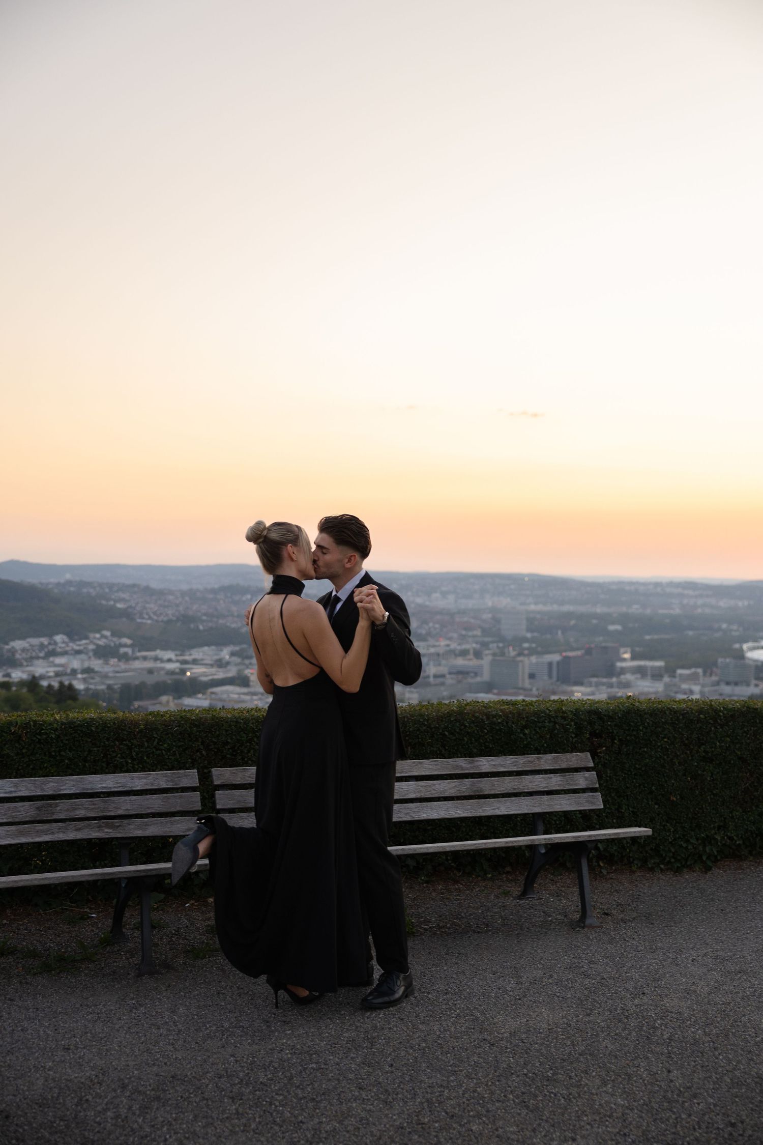 Elegant couple in black formal attire dancing at sunset with a scenic city view backdrop.
