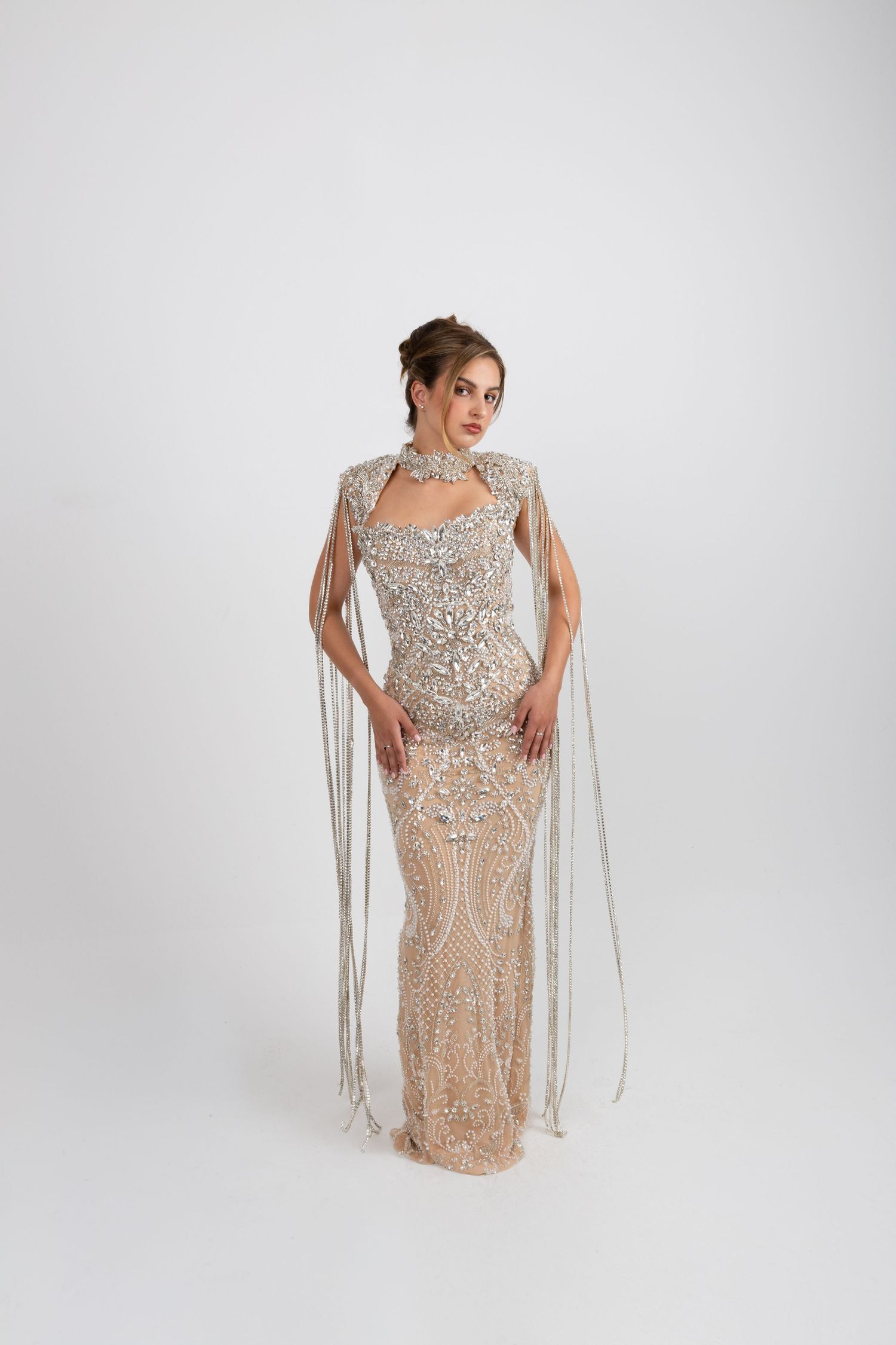 Woman in a floor-length beaded silver gown with flowing fringe cape sleeves and intricate embellishments on white background.