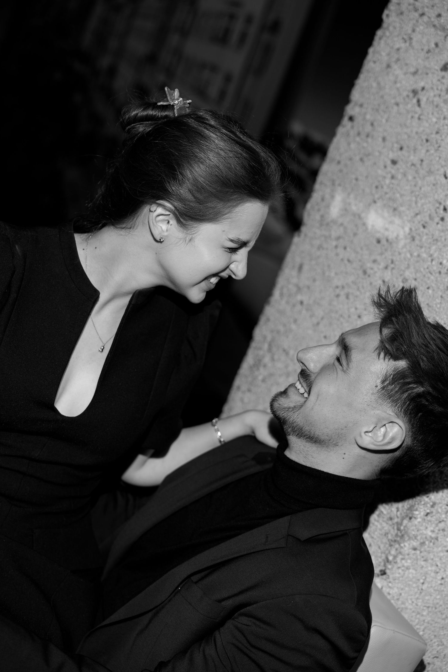 Black and white photo of laughing couple lying together, woman in black dress leaning over smiling man.