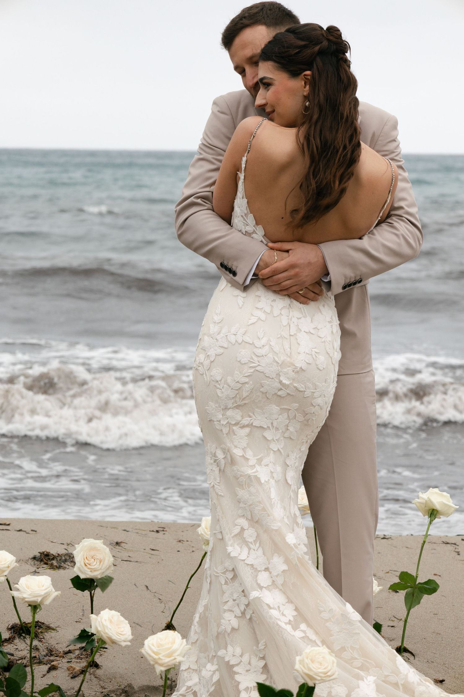 Bride in lace mermaid gown embraced by groom on a beach with white roses in foreground.