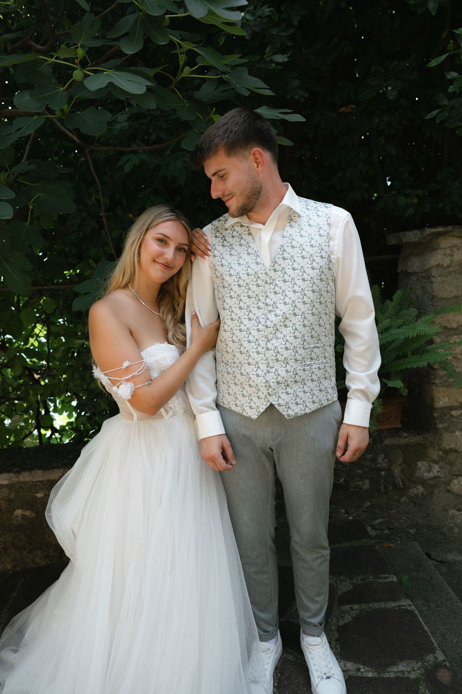 A smiling bride in a white tulle gown stands with a groom in a floral vest against lush green foliage.