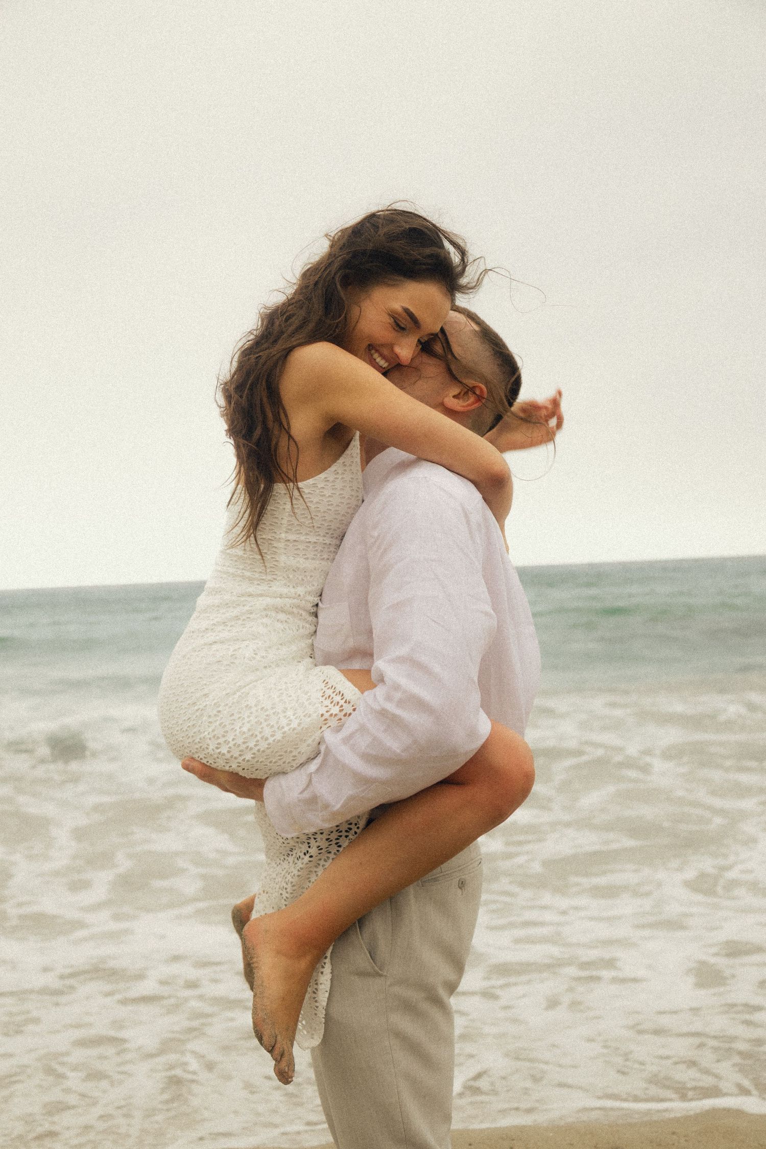 Romantic couple embracing on a beach, man lifting woman in white dress by ocean waves.