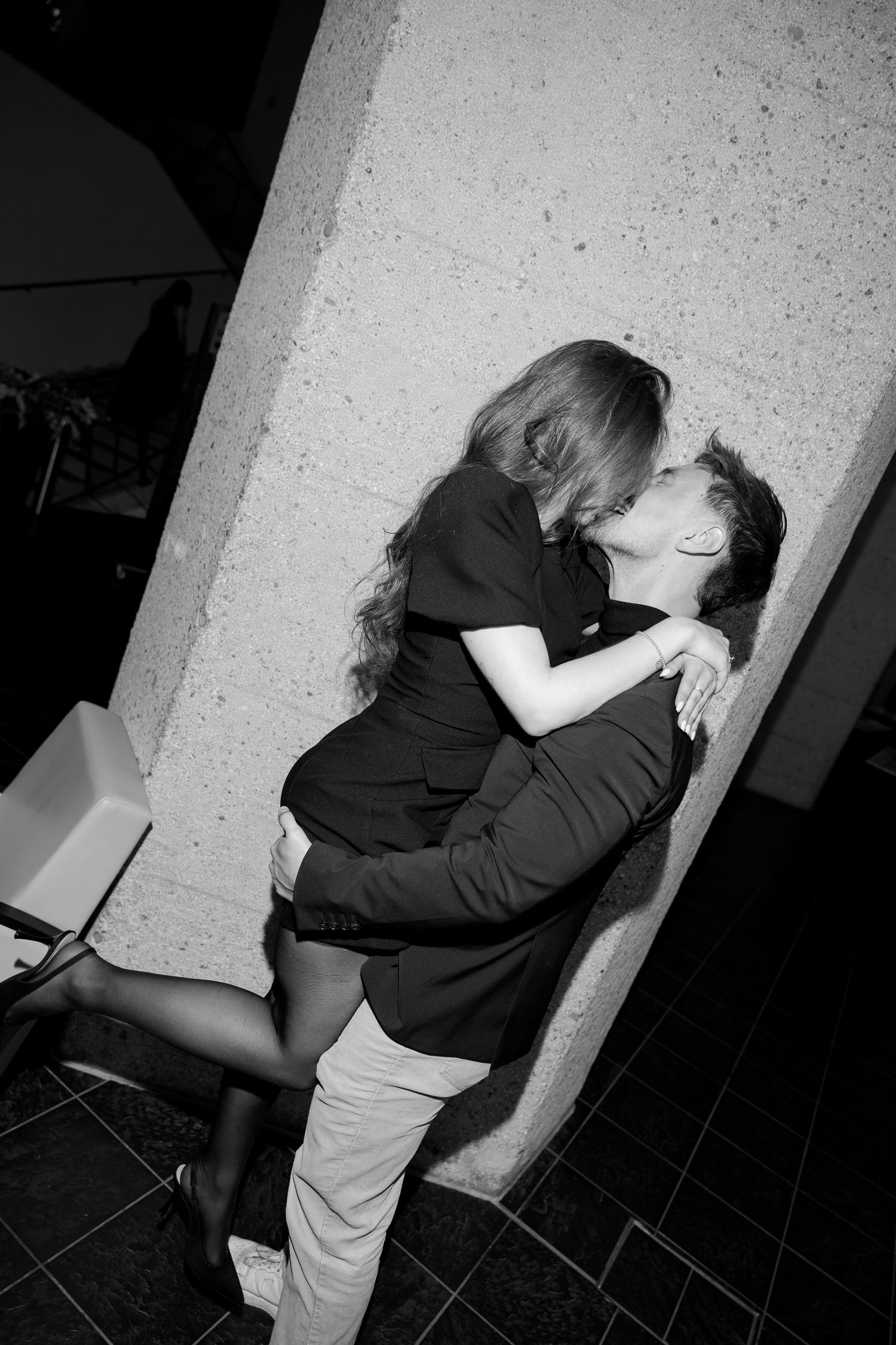 Black and white image of a couple kissing intimately against a light stone pillar indoors.