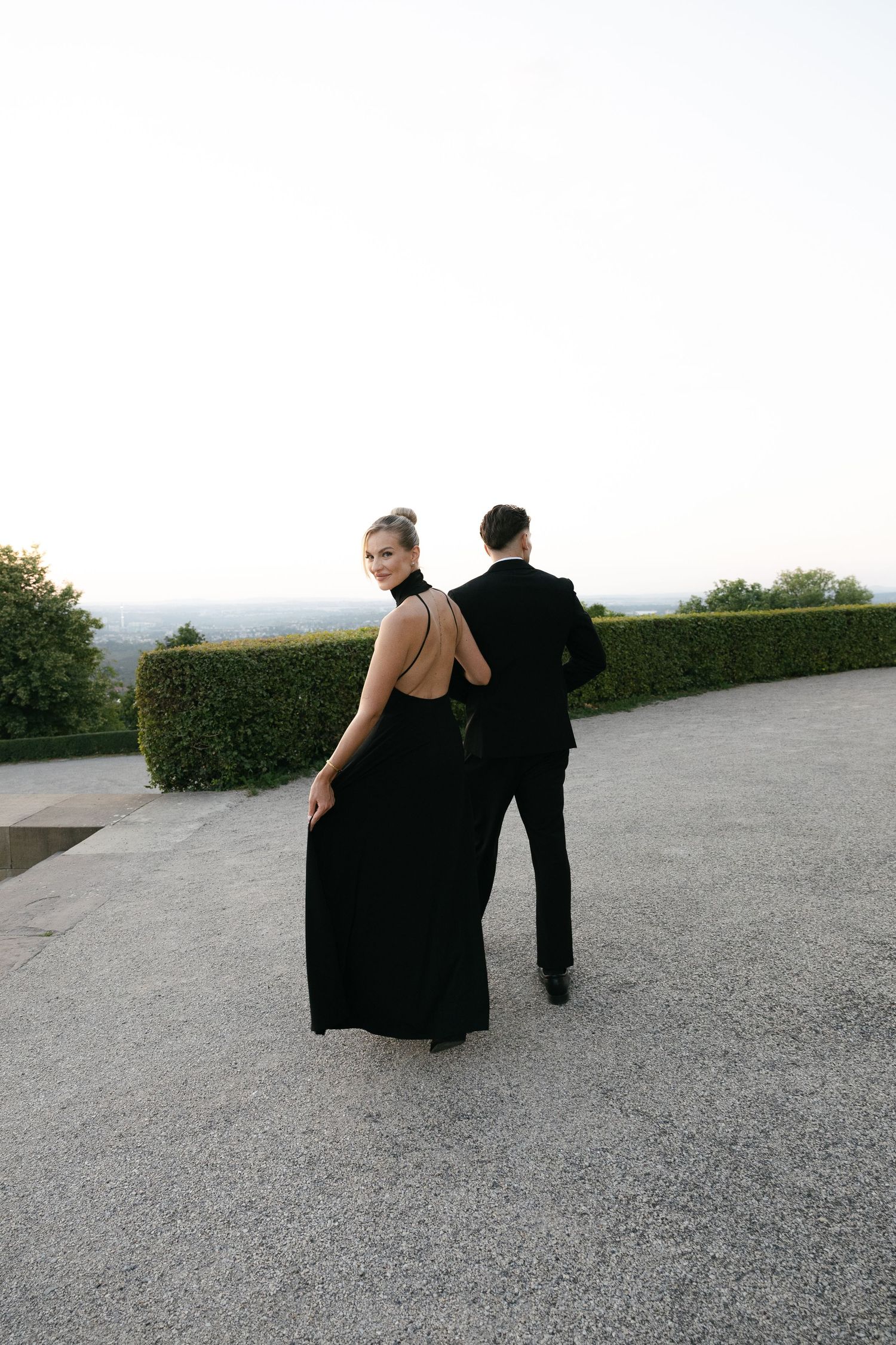 Elegant couple in black formal attire on a stone terrace, woman in backless gown glancing back, lush hedge backdrop.